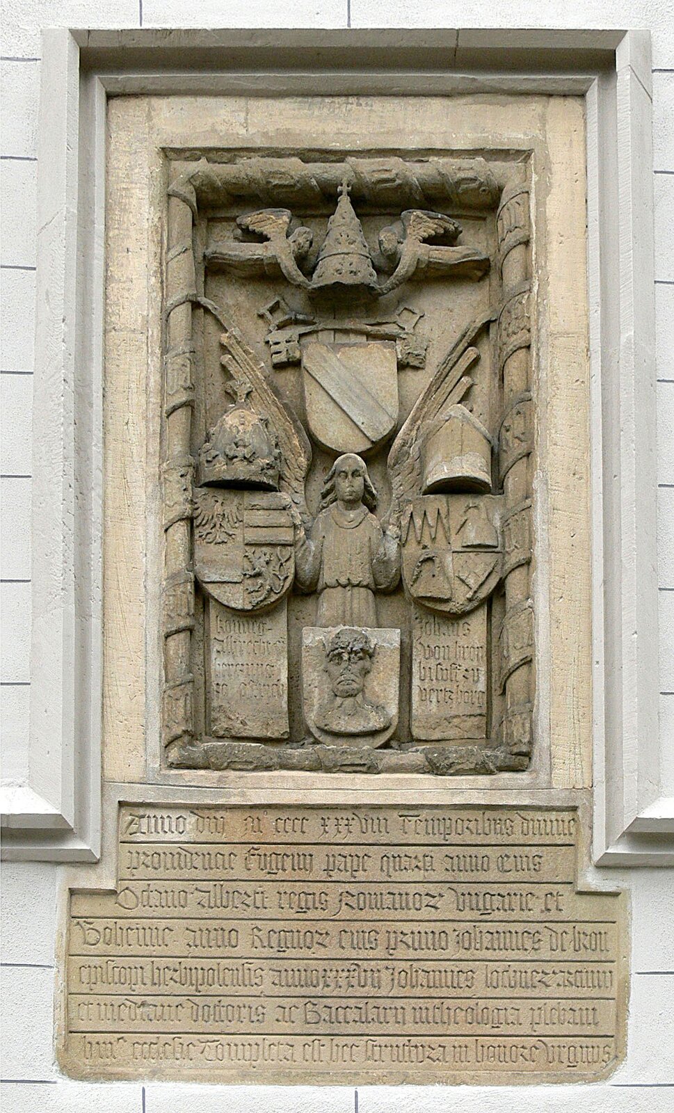 Zdjęcie przedstawia płaskorzeźbę na ścianie budynku. Na samej górze mira papieska trzymana przez dwóch aniołów. Pod nim tarcza z szarfą, a nad nią dwa skrzyżowane klucze. Pod herbem anioł opierający się o dwie tarcze. Tarcza po lewej ma orła i lwa, a nad tarczą korona króla rzymskiego. Po prawej tarcza, która nad sobą ma mirę biskupią. Pod tarczami napisy, a nogi anioła zakrywa tarcza z głową. Pod rzeźbą są napisy.