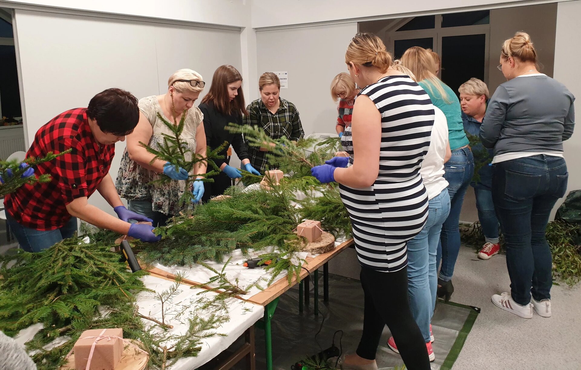 Na zdjęciu znajduje się grupa kobiet, która stoi przy stołach. Część osób trzyma w rękach gałązki z iglastego drzewa. Na stołach również znajdują się gałązki drzew, pistolety na gorący klej oraz podstawy do stroików kwiatowych. 