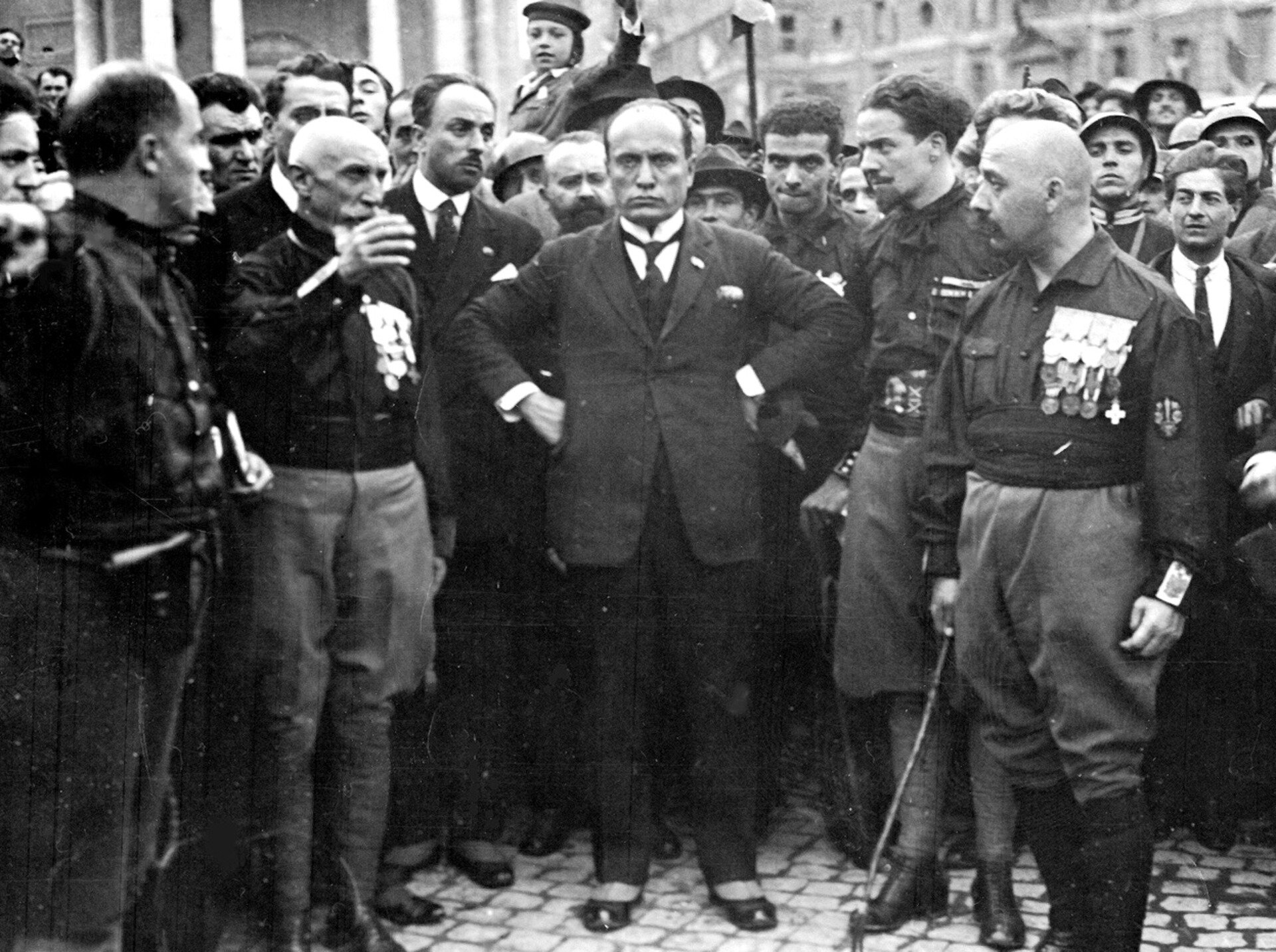 Fotografia czarno-biała przedstawiająca uczestników marszu w Rzymie. Pośrodku zdjęcia stoi Mussolini. Po lewej i prawej stronie otaczają go dowódcy wojskowi w odświętnych mundurach, a tle widoczni są zwykli żołnierze i obywatele.