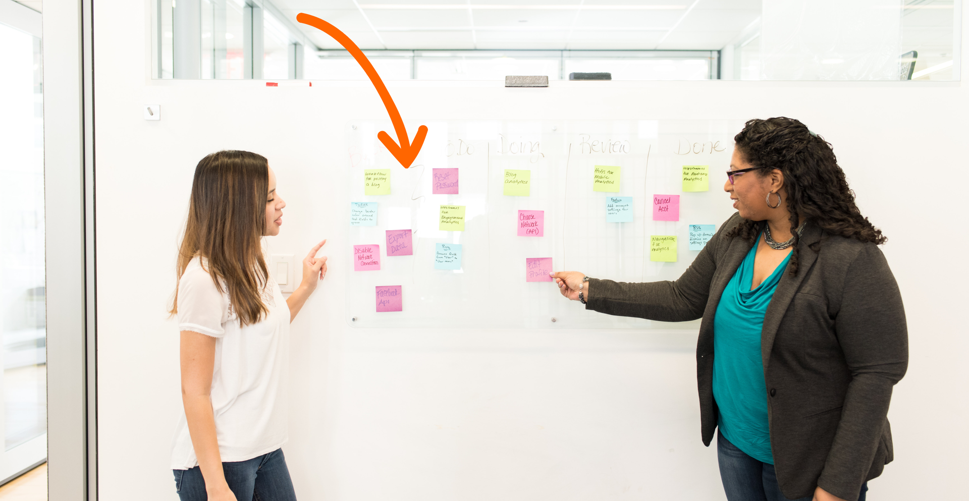 Zdjęcie przedstawia dwie kobiety stojące przy tablicy. Na tablicy przyczepione są małe karteczki. Strzałka na ilustracji skierowana jest na tablicę.