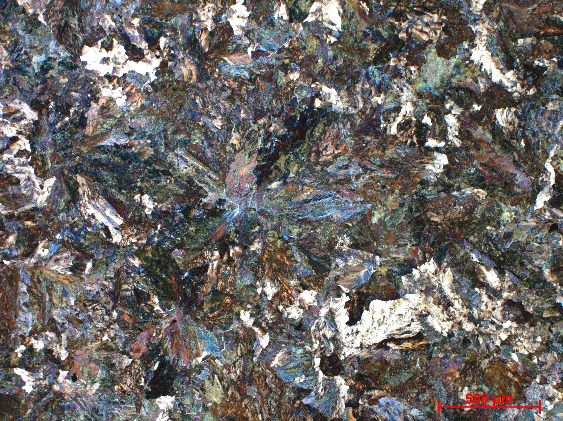 Zdjęcie przedstawia zdjęcie próbki stopu o zawartości węgla siedemdziesięciu siedmiu setnych procenta. Pokazana jest struktura perlityczna w powiększeniu pięciokrotnym. Widoczne są mieniące się różnymi kolorami struktury krystaliczne.