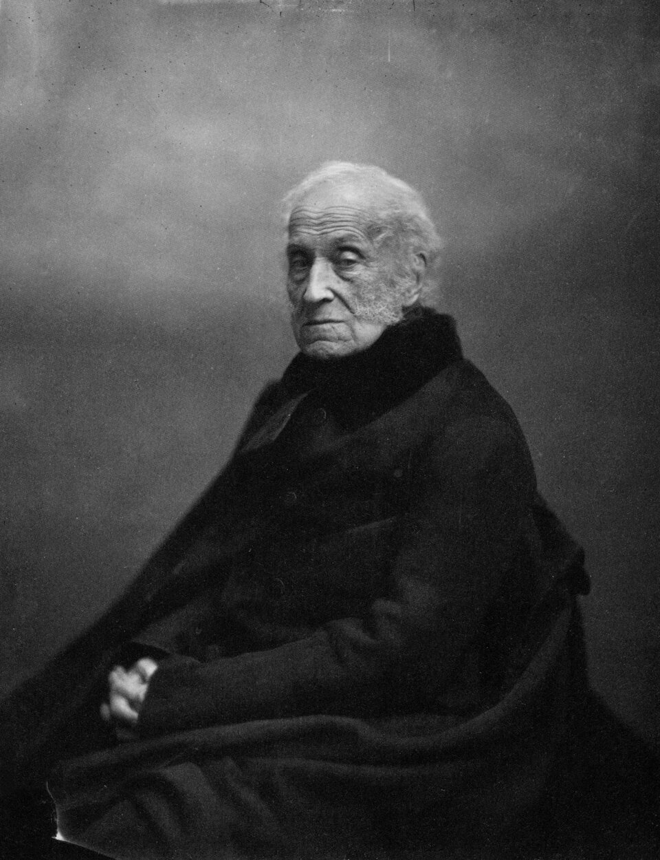 Biało‑czarny portret mężczyzny w podeszłym wieku. Ma lekką łysinę, siwe włosy, zarośnięte policzki. Skóra niczym rzeźba oddaje stateczność sytuacji. Ubrany na czarno być może kożuch. Ręce swobodnie opadają na kolona. Dłonie złożone niczym do modlitwy. 