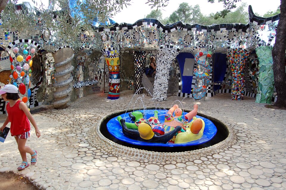 Ilustracja przedstawia pracę Niki de Saint Phalle pt. „Ogrodu tarota”. Dzieło pochodzi z 1980 roku. Ogród znajduje się w Capalbio w Toskanii we Włoszech. Na fotografii został przedstawiony element ogrodu. W centralnym punkcie znajduje się kolorowa, mała fontanna, w której umieszczone są figury postaci. W tle widoczne są kolumny budowli składające się z wielu mozaikowych elementów.