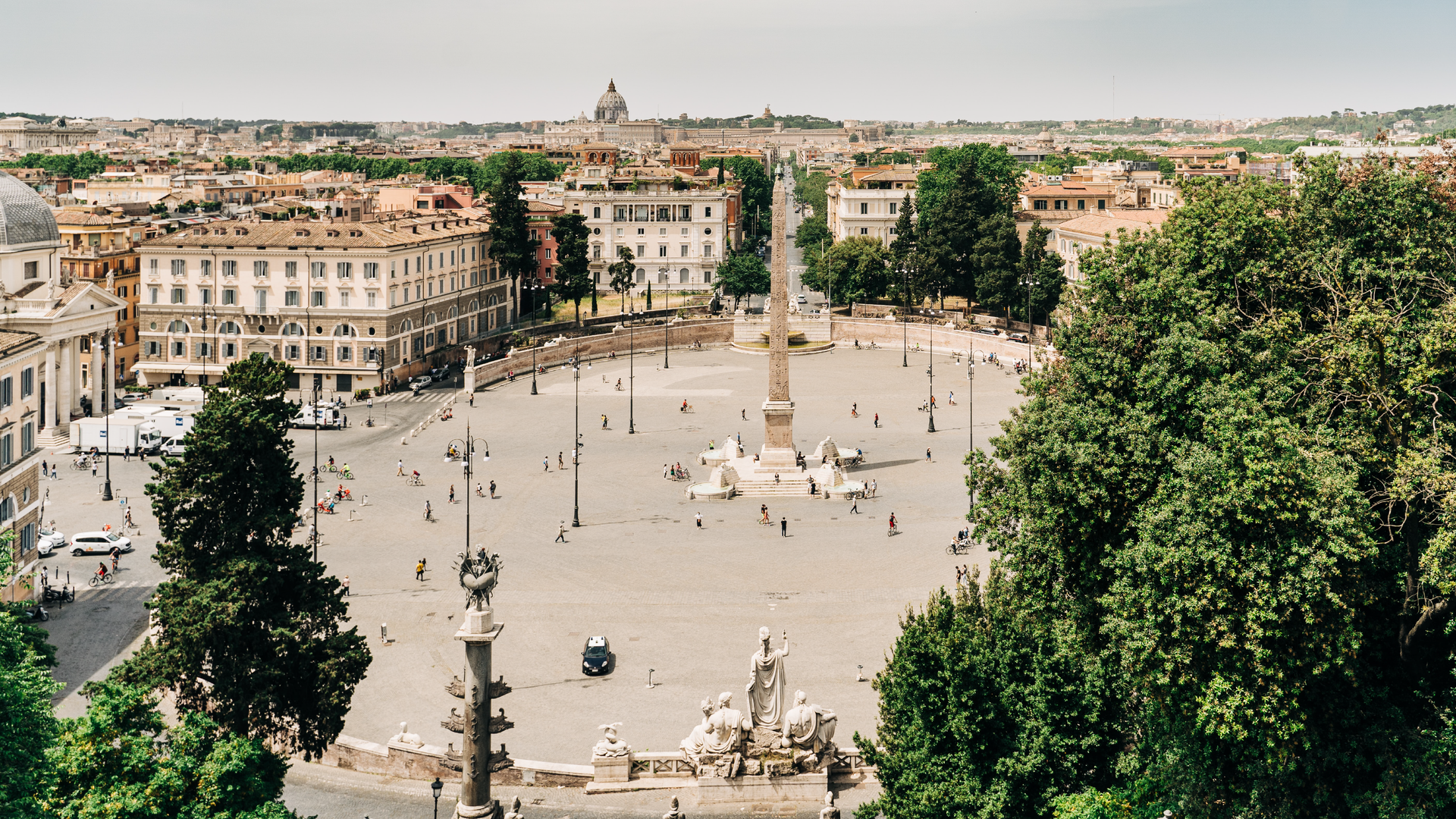 Zdjęcie przedstawia z góry Plac Ludowy w Rzymie. Plac jest okrągły, na środku znajduje się kolumna. Wokół placu widoczne są budynki i drzewa.