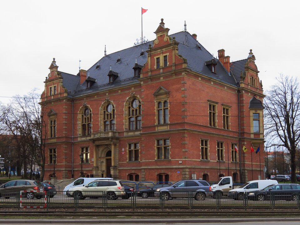 Na zdjęciu został przedstawiony czterokondygnacyjny budynek w stylu neorenesansowym wykonanym z cegły. Zdobiony jest gzymsami oraz boniami na krawędziach ścian. Wejście do budynku skonstruowane zostało w formie portalu z kolumnami po bokach. Na szczycie dachu widoczny jest masz i powiewająca na nim flagach w kolorze czerwonym. Przed budynkiem znajduje się ulica i jadące przez nią samochody oddzielone ogrodzeniem od torów tramwajowych.