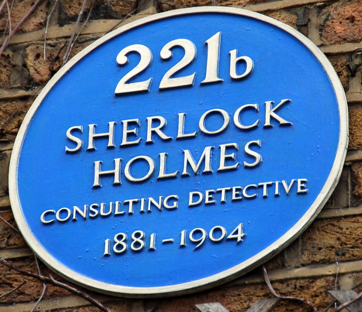 Na zdjęciu znajduje się szyld z napisem w języku angielskim na niebieskim tle: 221b Sherlock Holmes consulting detective 1881‑1904. W tle ceglana ściana i fragmenty gałęzi.