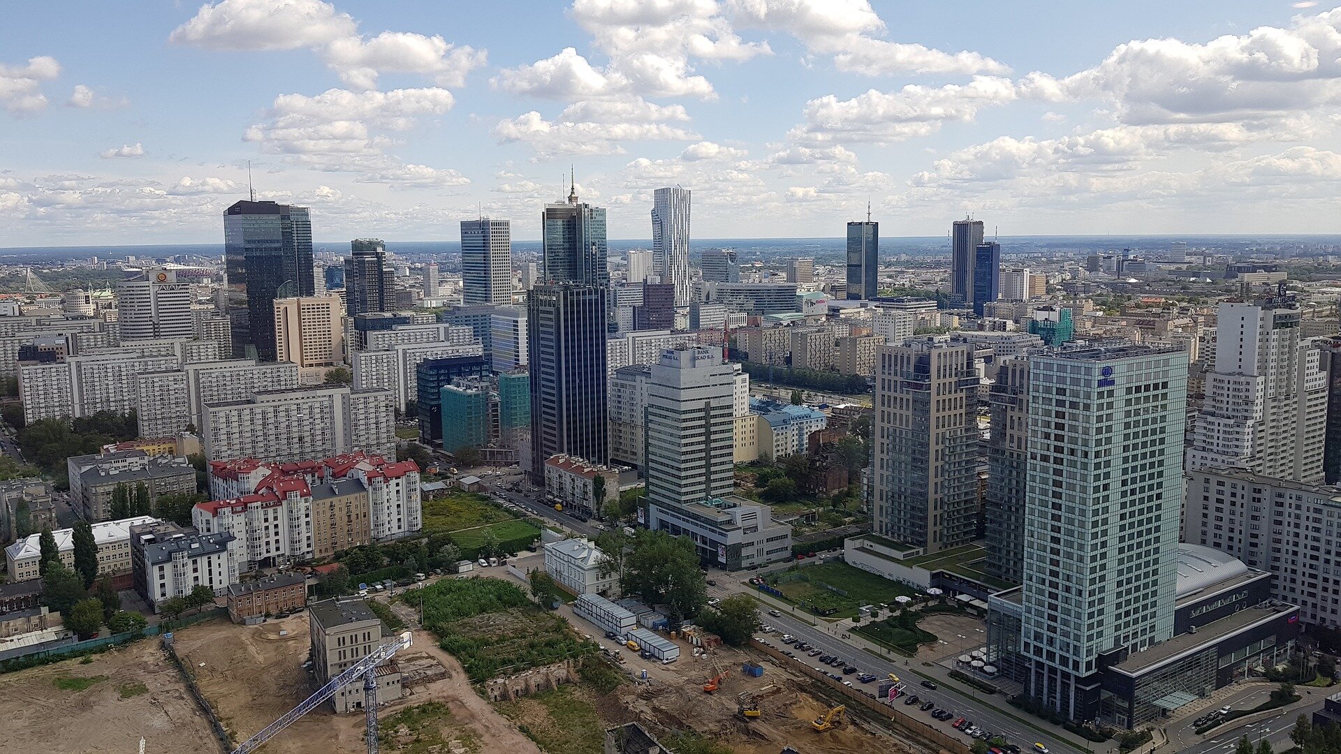 Ilustracja przedstawia zdjęcie z lotu ptaka na zabudowę w Warszawie. Widoczne są wysokie apartamentowce, biurowce. Na dole zdjęcia widać plac budowy, prawdopodobnie powstaną tam nowe budynki.