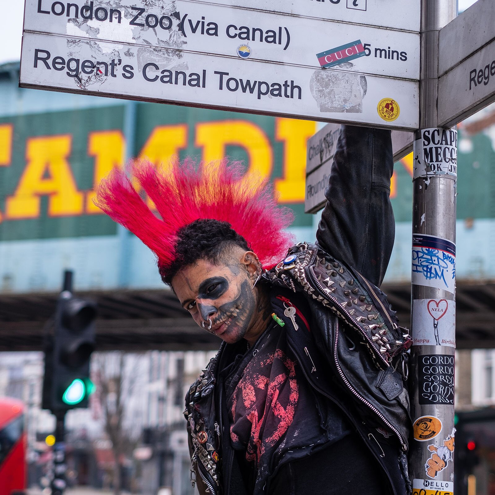 Zdjęcie przedstawia młodego mężczyznę, od pasa w górę, ubranego w czarną, skórzaną kurtkę nabijaną ćwiekami i czarną koszulkę. Włosy ma postawione w irokeza, w kolorze czerwonym. Na twarzy ma namalowany w czarnym kolorze szyderczy uśmiech i koła dookoła oczu. Lewą ręką trzyma drogowskaz z nazwami w języku angielskim London Zoo (via Canal), Regent's Canal Towpath. W tle widoczne rozmazane szyldy i budynki. 