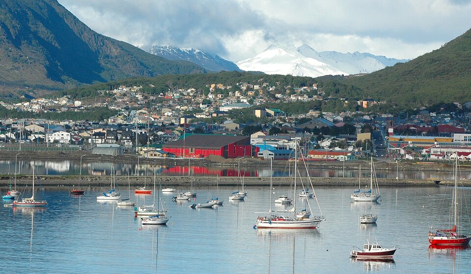 Zdjęcie przedstawia miasto położone nad brzegiem zbiornika wodnego. Na pierwszym planie jest woda i żaglówki. W tle są domy położone na zalesionym zboczu. W tle są ośnieżone górskie szczyty.  