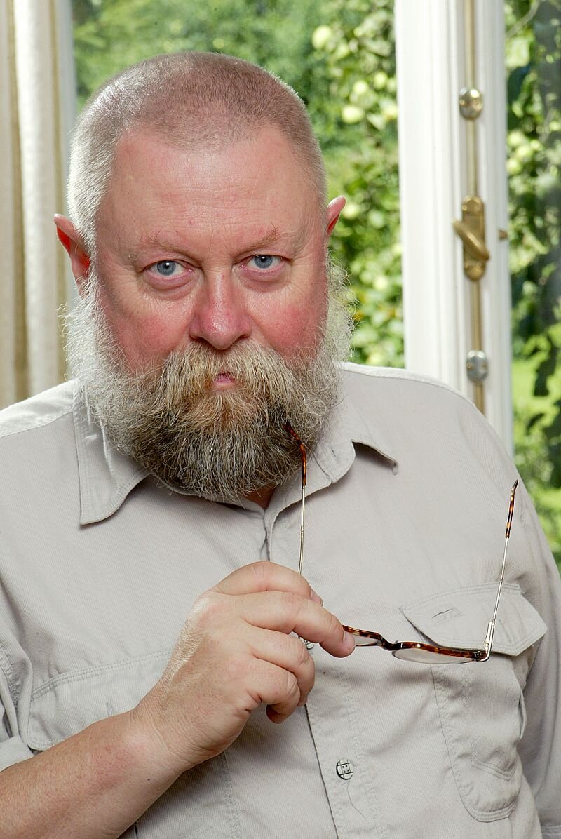 Zdjęcie przedstawia starszego mężczyznę. Jego twarz jest owalna, okolona gęstą brodą i wąsami. Mężczyzna ma na twarzy liczne zmarszczki. Jego nos jest wydatny, oczy podpuchnięte. Mężczyzna ma krótko ostrzyżone włosy. Ubrany jest w koszulę. z krótkim rękawem. W prawej dłoni trzyma okulary. Siedzi na tle okna, za którym znajduje się zieleń.