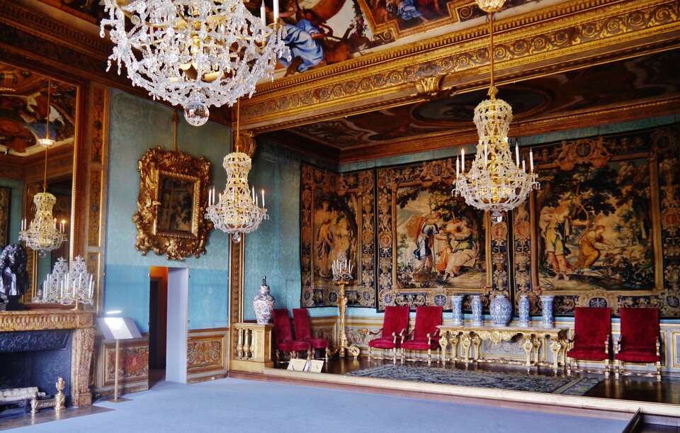 Ilustracja przedstawia wnętrze pałacu Vaux le Vicomte: „Salon Muz”. W kadrze znajduje się bogato zdobione wnętrze złoceniami, malowidłami sufitowymi, gobelinami i obrazami na ścianach, kryształowymi żyrandolami, lustrem nad kominkiem.