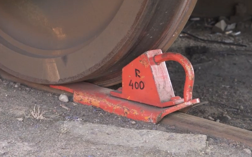 Zdjęcie przedstawia płozę hamulcową. Jest to podłużny kawałek metalu z klinem i uchwytem, położony na szynie i blokujący koło. Pomalowana jest na czerwono, a na boku klinu znajduje się podpis z numerem posterunku manewrowego do którego należy płoza.