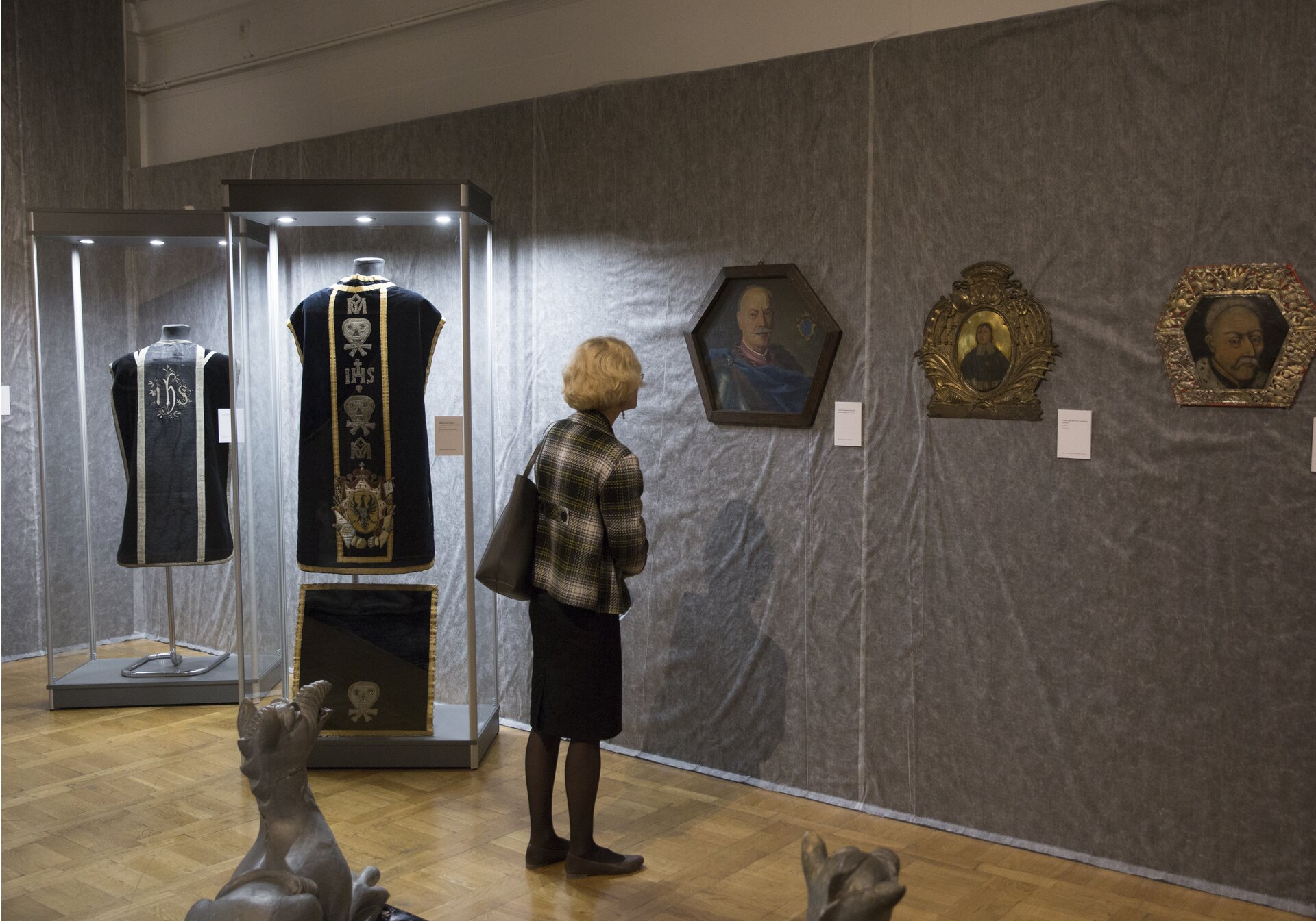 Zdjęcie ukazuje wnętrze galerii lub sali muzealnej gdzie prezentowana jest kolekcja dzieł sztuki. Na szarej ścianie wiszą trzy obrazy, przed którymi stoi kobieta ubrana w czarną spódnicę oraz marynarkę w kratę. Z lewej strony, nieopodal kobiety, w gablotach wiszą dwa czarne zestawy ubrań. Pierwsze, wiszące w gablocie, na stojaku to czarna szata zdobiona złotymi paskami materiału, pomiędzy którymi umieszczono srebrne napisy IHS oraz symbole czaszek i kości. U dołu szaty umieszczony został herb. Niżej pod szatą opiera się o nogę stojaka, materiał w kolorze czarnym. Jest on zdobiony złotymi paskami materiału. U dołu także widnieje symbol czaszki i kości. Stojąca w drugiej gablocie szata również jest czarna. Zdobiony jest srebrnymi paskami materiału pomiędzy którymi użyto szarego barwnika oraz białego napisu IHS, gdzie każda z liter ozdobiona została ornamentem. 