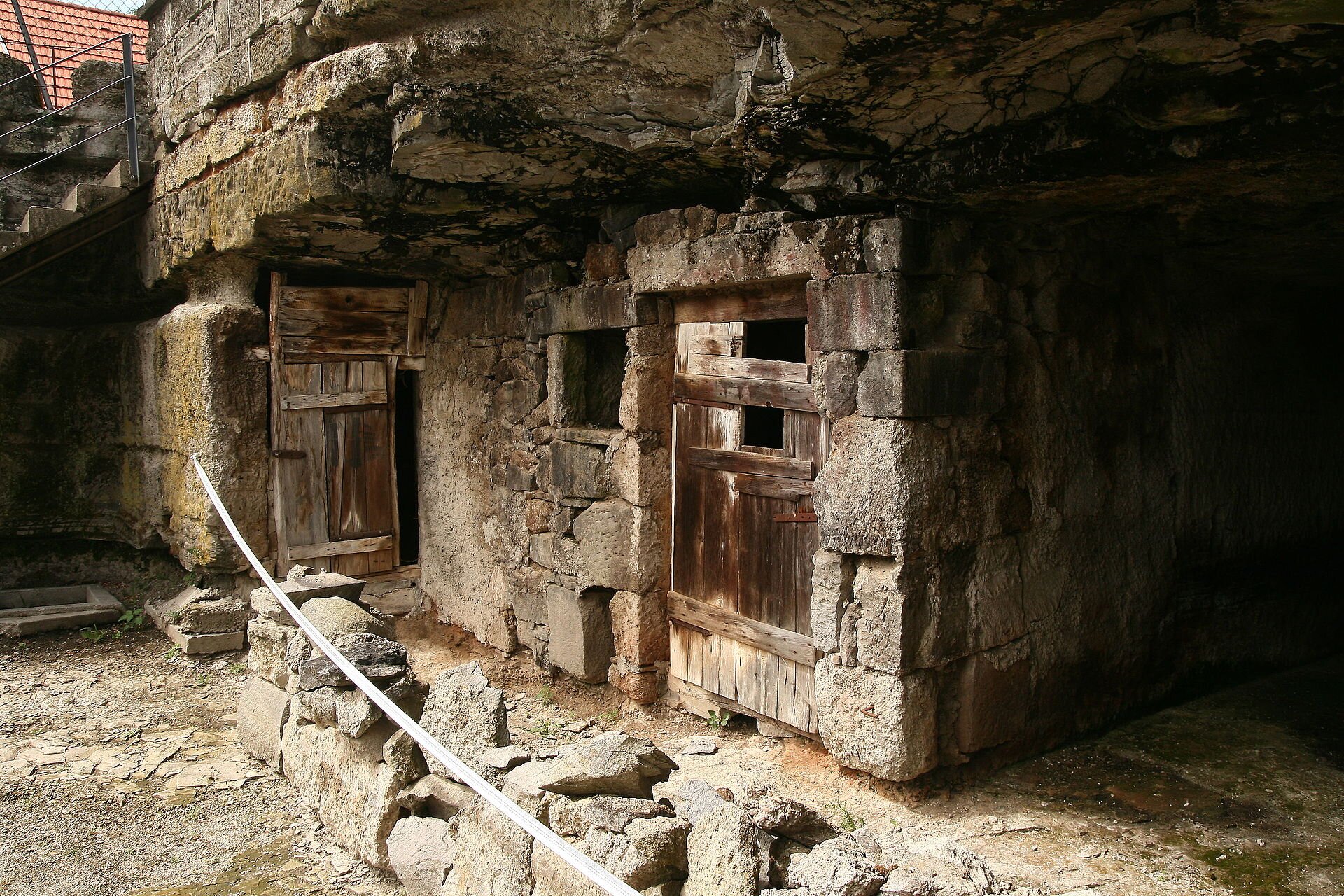 Fotografia domu wykutego w skale. Prowadzą do niego drewniane drzwi. Przed nim kamienny murek.