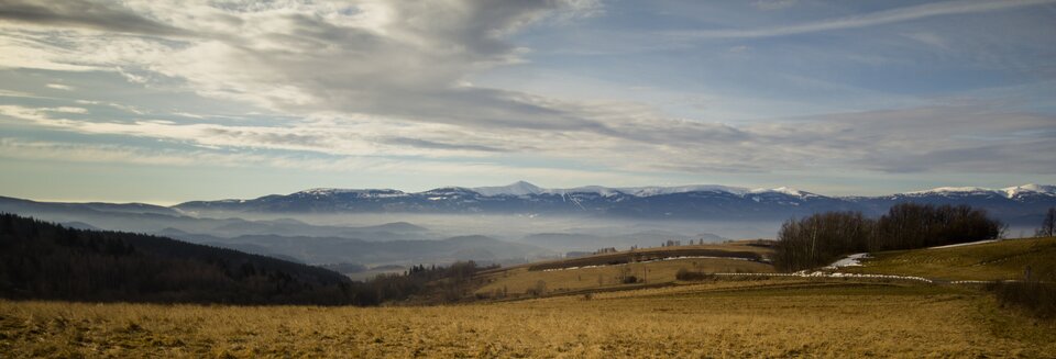 Na zdjęciu znajduje się panorama łagodnych pofalowanych terenów porosniętych suchą trawą. Gdzieniegdzie rosną drzewa liściaste w szacie jesiennej. Dalej widoczny jest niżej położony teren zasnuty mgłą. Na linii horyzontu znajduje się długie pasmo górskie ze szczytami pokrytymi śniegiem. Ponad górami niebo jest dość jasne, ze smugami chmur. 