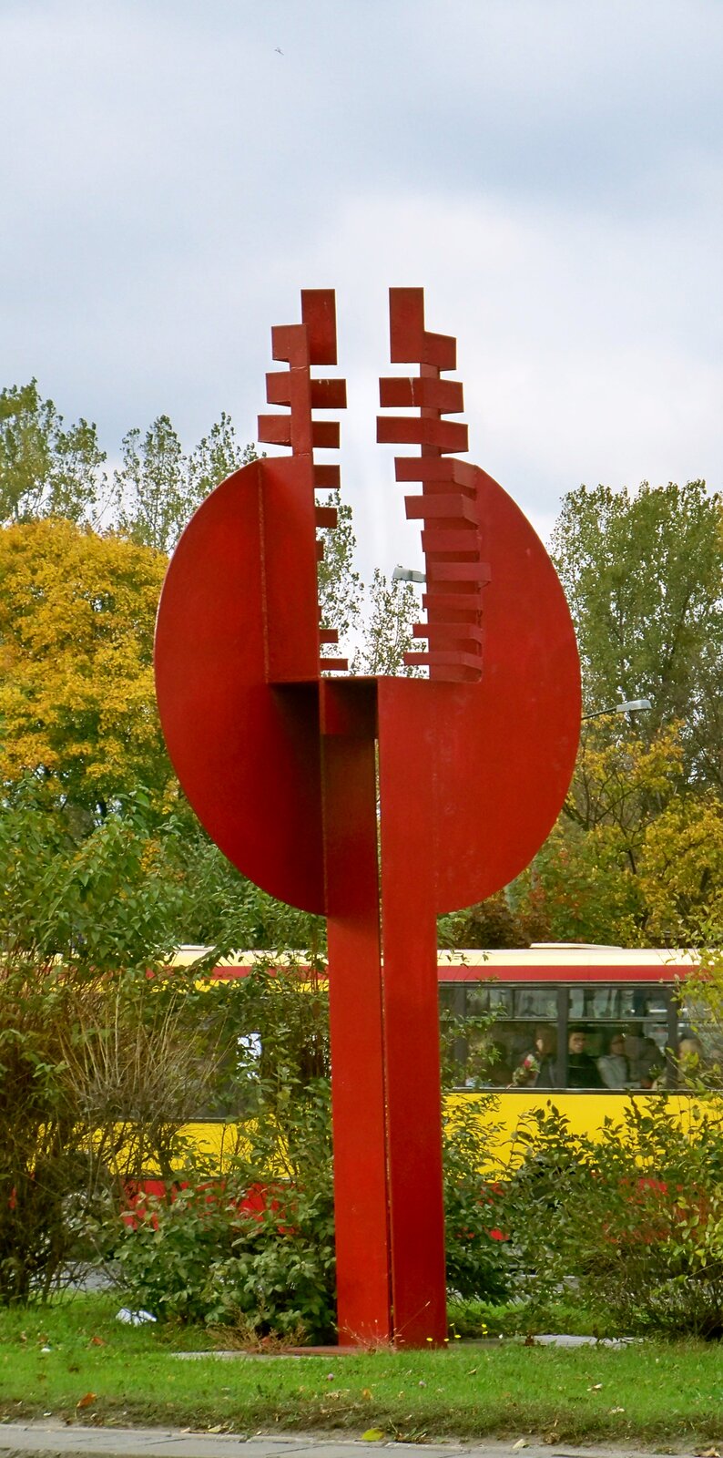 Fotografia przedstawia rzeźbę Macieja Szańkowskiego, po tytułem: „Biennal”. Wykonana z metalu, geometryczna, intensywnie różowa rzeźba, stoi w plenerze wśród zielonych krzewów. Na drugim planie w tle znajduje się autobus. 