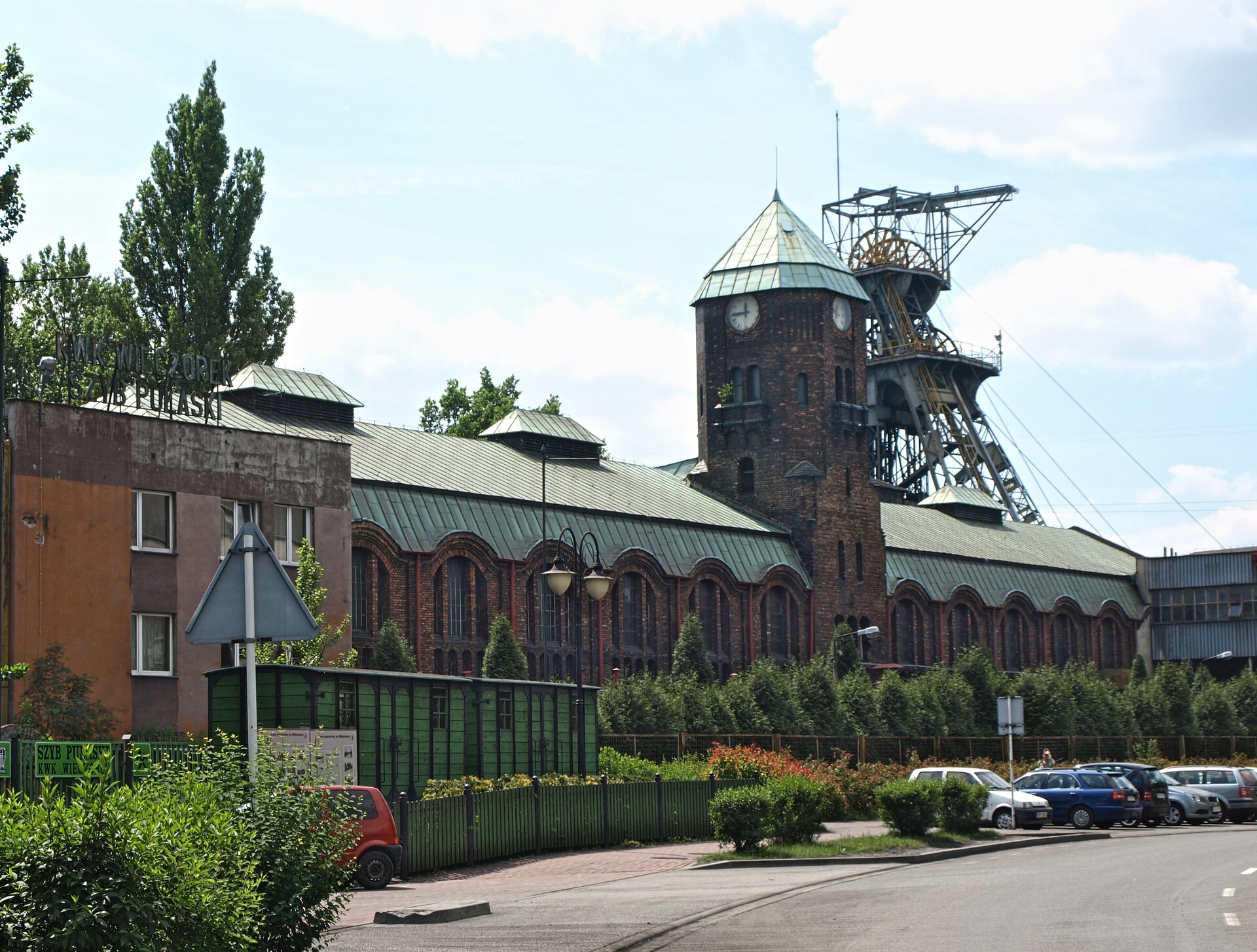 Na zdjęciu ceglany budynek z wieżą. W tle wieża szybowa – wysoka konstrukcja stalowa z kołami. Przed budynkiem zaparkowane samochody.