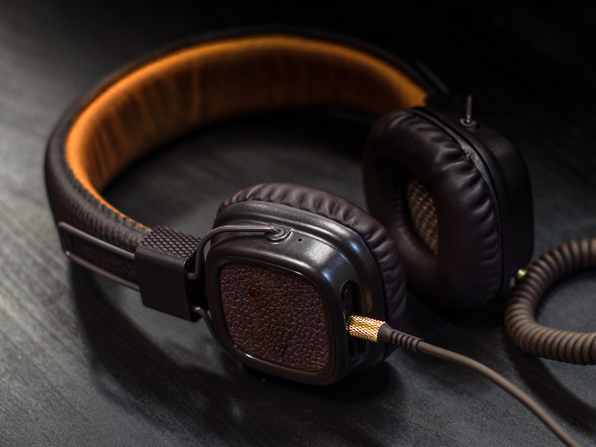 Kolorowa fotografia przestawia przewodowe słuchawki nauszne. Kwadratowe, czarne głośniki połączone są grubym pałąkiem, wyścielonym od wewnątrz pomarańczową gąbką.