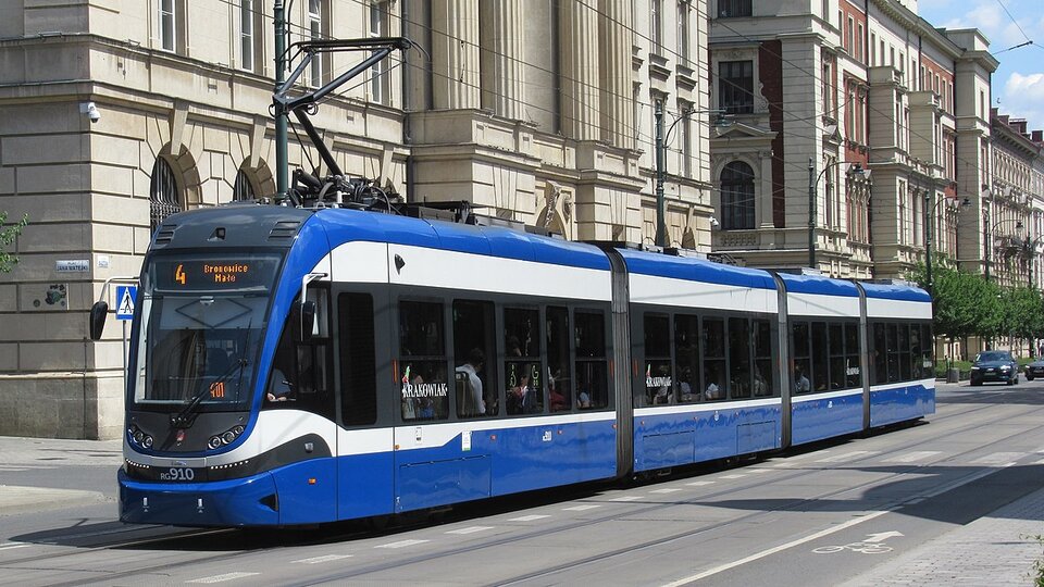 Na zdjęciu widać tramwaj jadący po torowisku, pas rowerowy przy krawędzi jezdni oraz pierzeję kamienic w tle. Za tramwajem jadą samochody.