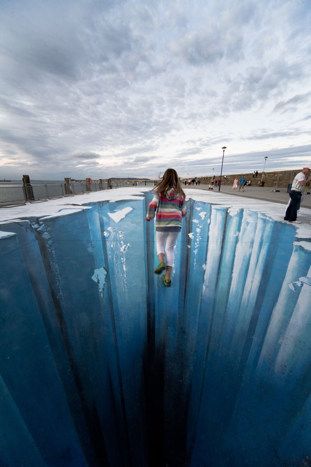 Zdjęcie przedstawia złudzenie optyczne namalowane na molo, po którym biegnie mała dziewczynka, ubrana w białe spodnie oraz bluzę w wielokolorowe paski. Obraz 3D prezentuje przepaść w lodowcu, którego fragmenty odpadają i kruszą się, spadając w otchłań. Dziewczynka również wygląda jakby wpadała w otchłań.