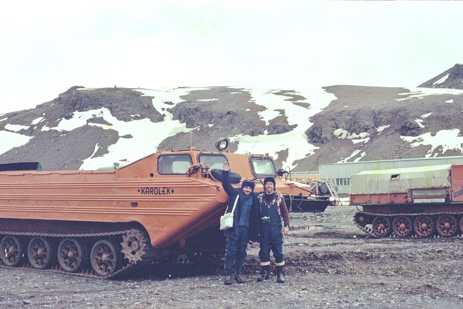 Zdjęcie przedstawia dwóch obejmujących się mężczyzn, pozujących do zdjęcia. Za nimi pojazdy na gąsienicach, a dalej lekko ośnieżone wzniesienia.