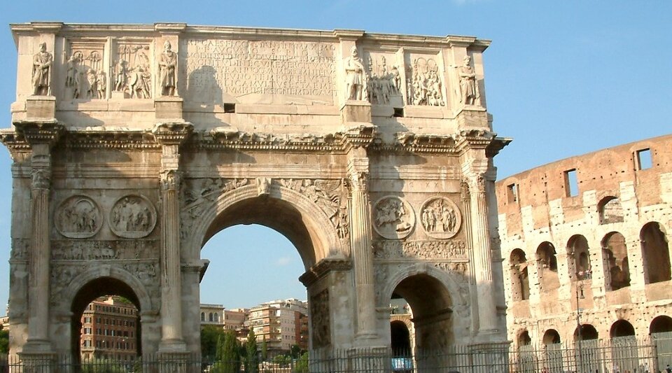 Zdjęcie ukazuje łuk triumfalny. To budowla w kształcie monumentalnej, wolno stojącej bramy. Ma liczne zdobienia. Po prawej stronie jest fragment okrągłego budynku z licznymi arkadami. 