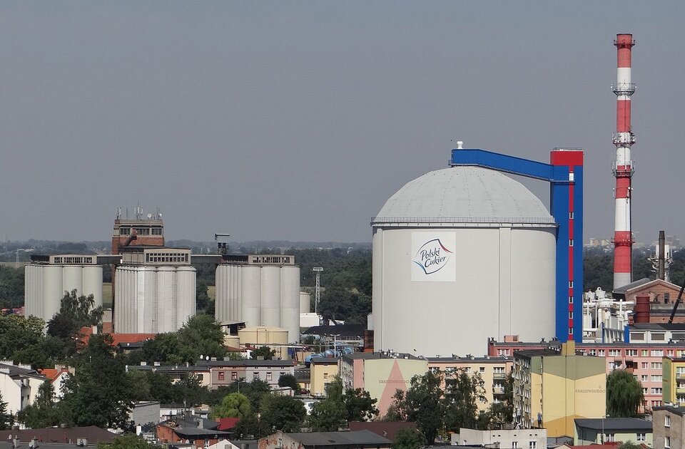 Zdjęcie przedstawia budynki cukrowni. Po prawej stronie jest duży, okrągły silos i komin. Po lewej stronie są niższe silosy.  