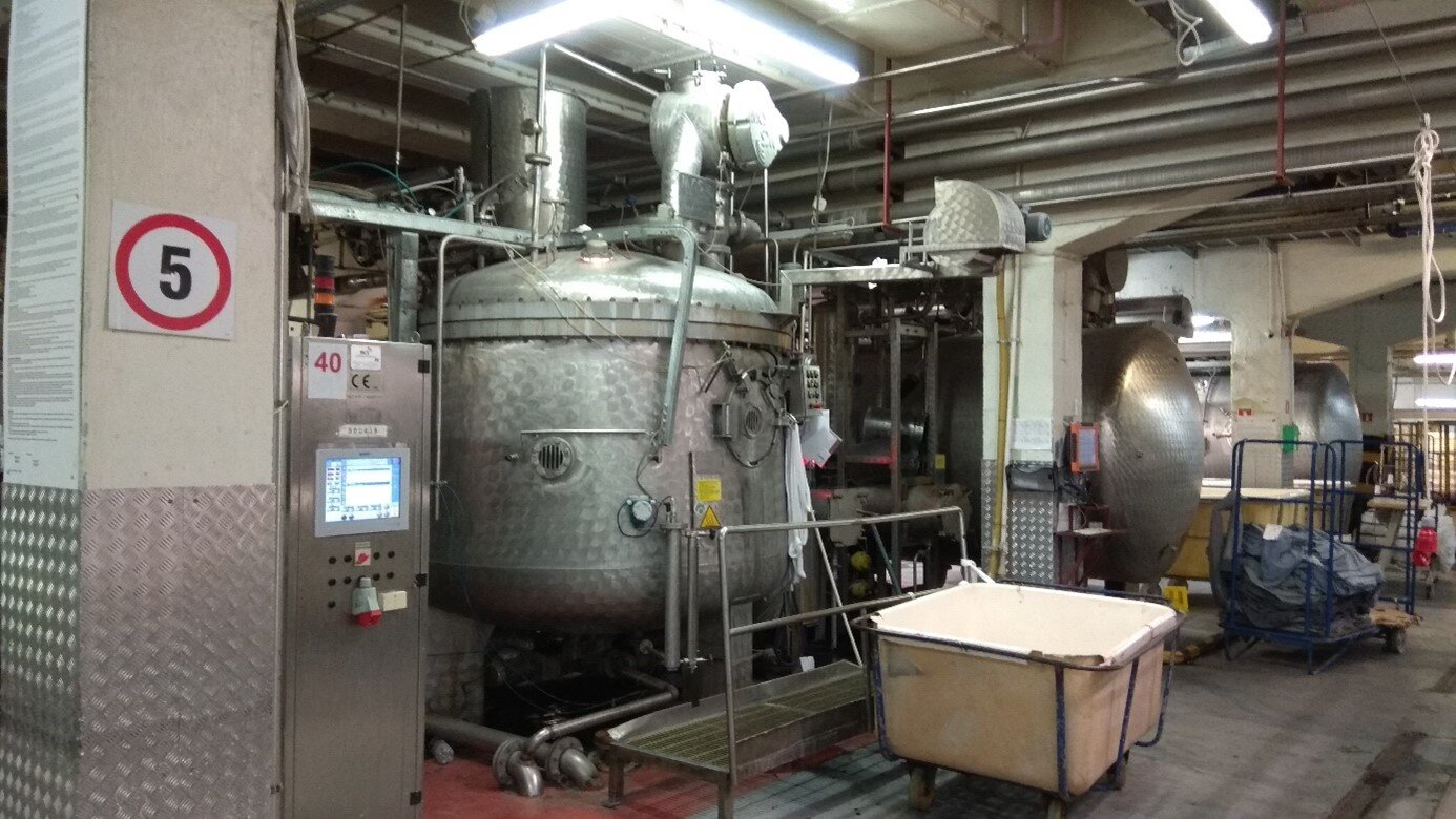 Zdjęcie przedstawia napawarkę stojącą w hali produkcyjnej. Metalowe urządzenie składa się z dużego, walcowatego zbiornika oraz licznych rur. W tle dwa inne zbiorniki oraz wózki na dzianiny.