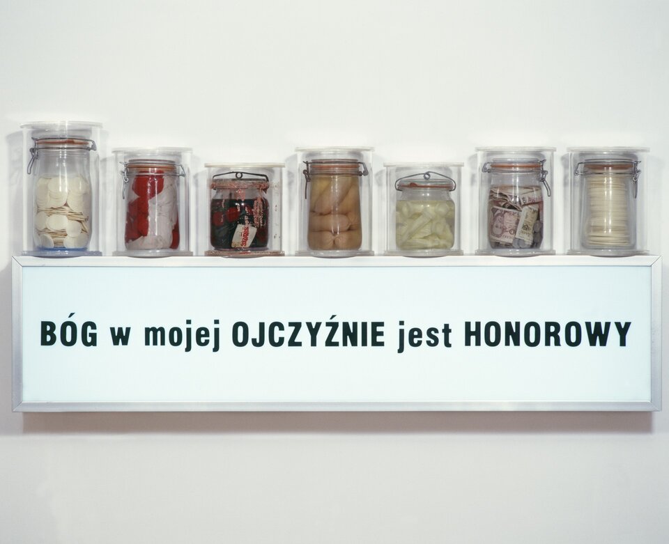 Ilustracja przedstawia obraz Roberta Rumasa „Bóg w mojej ojczyźnie jest honorowy”. Na fotografii widzimy siedem szklanych słoików o różnych rozmiarach. Ustawione są one w jednym rzędzie. W każdym słoiku znajduje się inne wypełnienie. Są to m.in. pieniądze, parówki, a także opłatek. Słoiki ustawione są na półce, pod którą widnieje napis BÓG w mojej OJCZYŹNIE jest HONOROWY. Autorem pracy jest polski artysta wizualny i kurator sztuki związany z nurtem sztuki krytycznej.