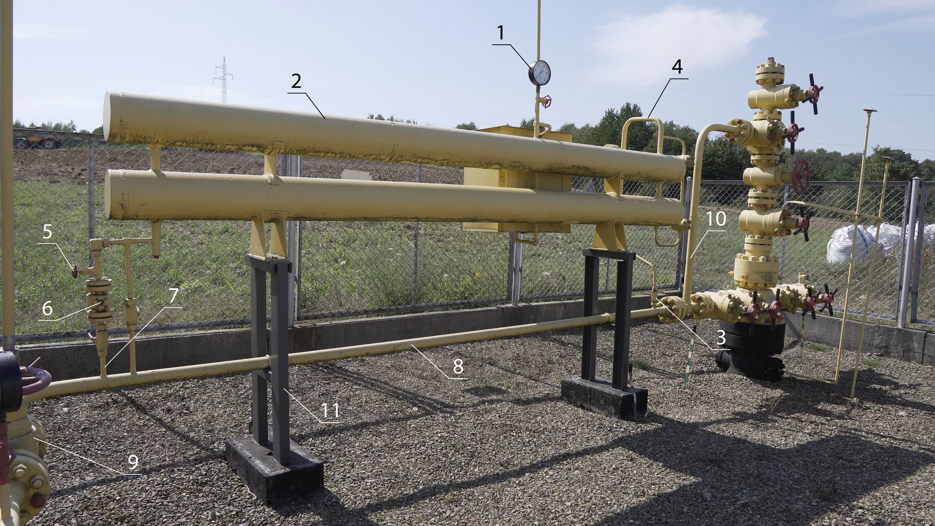 Na rysunku widoczne jest zdjęcie przyodwiertowego zakraplacza metanolu wykonane pod kątem. Składa się on z dwóch cylindrów koloru żółtego, wydłużonych w poziomie połączonych ze sobą dwoma pionowymi poprzeczkami po zewnętrznej stronie i łącznikami konstrukcyjnymi na szerokości filarów podtrzymujących konstrukcję Górny cylinder oznaczony jest cyfrą 3 oznaczającą zawór kulowy. Na szerokości wewnętrznych poprzeczek, pod dolnym cylindrem, widoczne są podwójne filary koloru czarnego które prowadzą do fundamentów oznaczone cyfrą 12. Pomiędzy podwójnymi filarami przechodzi rura koloru żółtego o małej średnicy oznaczona po środku cyfrą 9 jako zasuwa kolektora. Z lewej strony rura ta prowadzi do rozszerzenia usytuowanego pod kątem prostym do niej, składającego się z dwóch kołnierzy połączonych śrubami oraz od góry zawory z kołem regulacyjnym koloru czerwonego, od którego odchodzi kolanko w dół aż do ziemi.  Element ten oznaczony jest cyfrą 10 oznaczającą wlot gazu ziemnego do kolektora. Do elementu o kolorze żółtym dołączony jest okrągły element koloru czarnego zwrócony w przeciwną stronę do kierunku wykonywania zdjęcia. Z lewej strony rury małej średnicy koloru żółtego w górę odchodzą dwie cienkie rurki – lewa o widocznym znacznym rozszerzeniu w środku oznaczonym cyfrą 7 wskazującą na połączenie do dawkowania metanolu do kolektora, nad którym widoczna jest dalsza część pionowej rurki z ślepym odgałęzieniem w lewą stronę oznaczonym cyfrą 6 jako wziernik. Prawa pionowa rurka posiada w 2/3 wysokości niewielkie rozszerzenie oznaczone cyfrą 4 jako połączenie wyrównujące ciśnienie pomiędzy zakraplaczem a kolektorem gazowym. Obie cienkie pionowe rurki łączą się u góry z cienką rurką poziomą która łączy się z szerszą rurką pionową, ślepo zakończoną od dołu, a od góry połączoną z dolnym cylindrem. Na drugim planie za kołem wlotu gazu ziemnego do kolektora widoczna jest pionowa żółta rura wychodząca poza kadr zdjęcia. W połowie szerokości dwóch cylindrów za nimi widoczne jest prostokątny element koloru żółtego od którego w osi w górę wychodzi cienka rurka koloru żółtego z widocznym po środku okrągłym wskaźnikiem oznaczonym cyfrą 1 jako manometr. Z prawej strony cylindry są połączone rurą koloru żółtego z kolankiem oznaczonym cyfrą 5 jako zawór iglicowy. Pozioma rurka oznaczona cyfrą 9 jako zasuwa kolektorowa łączy się z prawej strony z rozszerzeniami stanowiącymi połączenia kołnierzowe ze śrubami. Przy pierwszym połączeniu widoczna jest rura pionowa koloru żółtego prowadząca kolankiem w prawo do głowicy oznaczona cyfrą 11 jako podstawa do zakraplacza. Rura ta łączy się z głowicą eksploatacyjną z prawej strony. W tle widoczne ogrodzenie oraz trawnik.