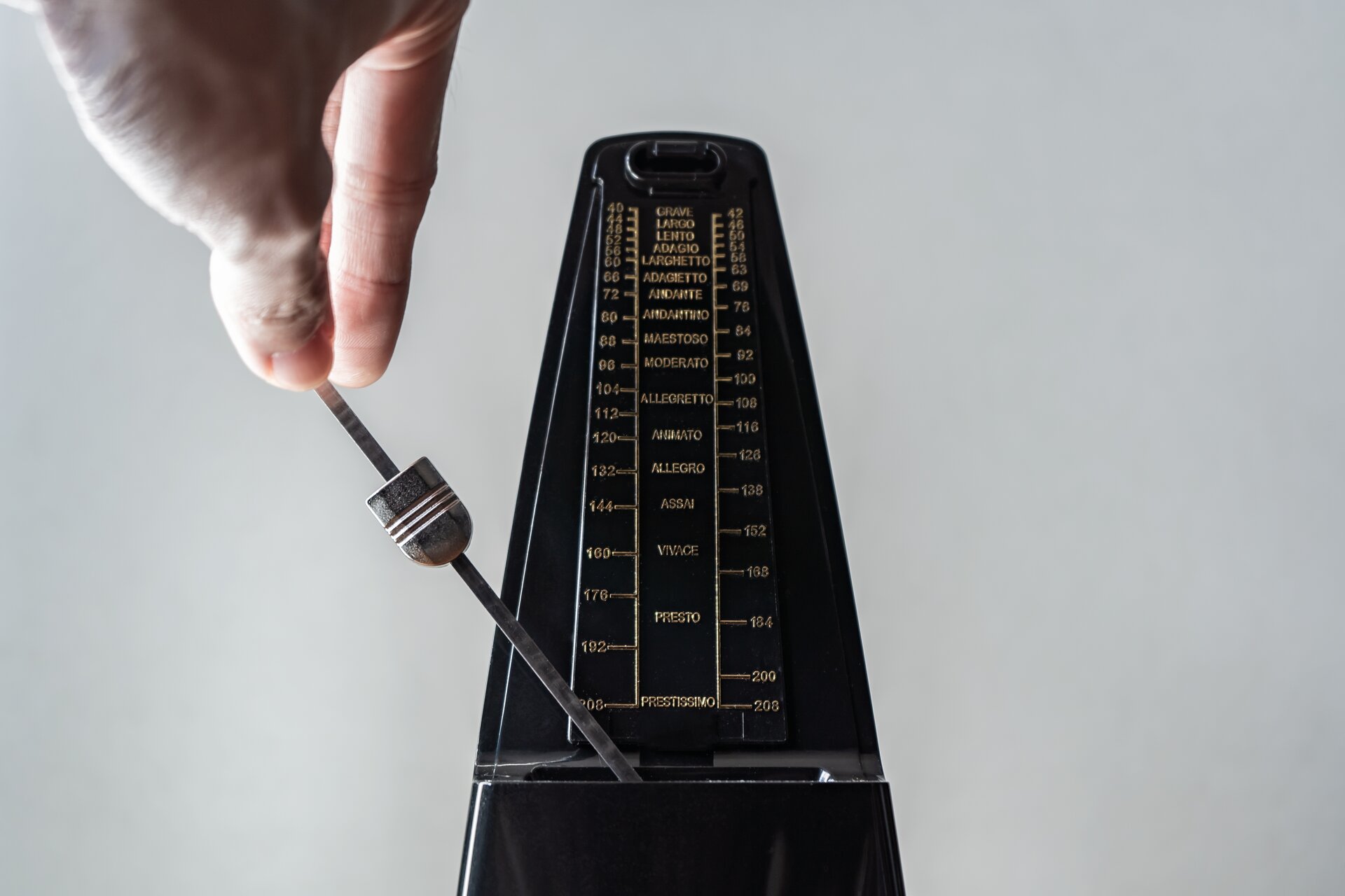 Na zdjęciu, na białym tle ściany zaprezentowane zostało urządzenie służące do podawania tempa utworów muzycznych. Przyrząd jest w kolorze czarnym, o kształcie zbliżonym do prostokąta, jednak patrząc od dołu do góry przedmiot zaczyna się zwężać. Są na nim napisane w złotym kolorze fachowe wskaźniki, a z samego przodu zamontowane jest wahadło, które kiwa się od lewej strony do prawej. Na tym cienkim fragmencie umieszczony został element, który można przesuwać od góry do dołu, tym samym przyspieszając bądź zwalniając jego ruch.