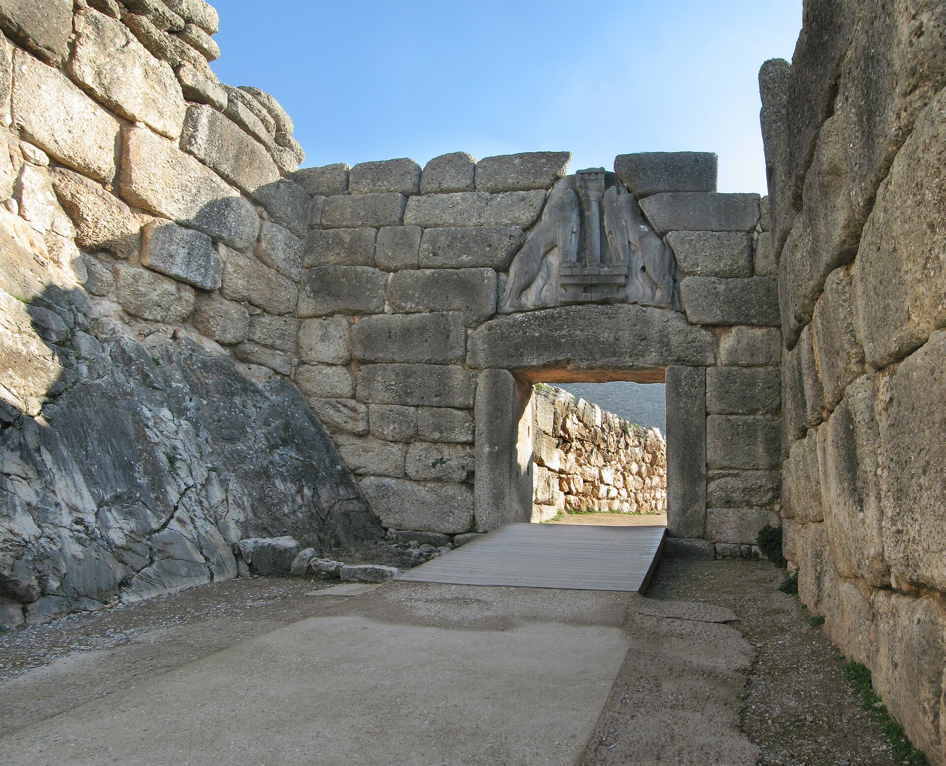 Na zdjęciu kamienna brama w murze. Nad otworem wejściowym trójkątna płyta z płaskorzeźbą z lwami.