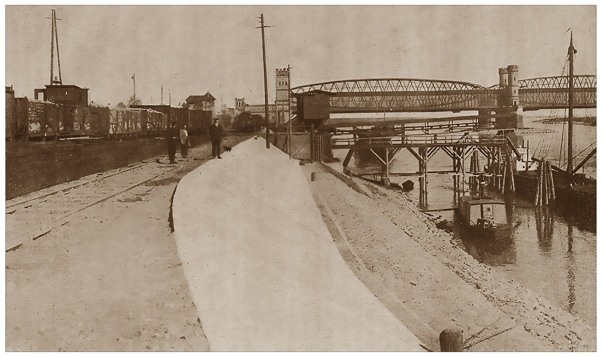 Zdjęcie przedstawia port rzeczny od strony północnej. Z prawej strony zdjęcia znajduje się wał przeciwpowodziowy, u dołu którego płynie rzeka. Przy brzegu znajdują się drewniane mosty z przycumowanym statkiem. Po lewej stronie zdjęcia, obok wału, deptak, po którym przechadzają się ludzie. Dalej, z lewej strony, tory, na których stoi pociąg. W tle, na rzece, widoczny jest murowany most łączący oba brzegi.