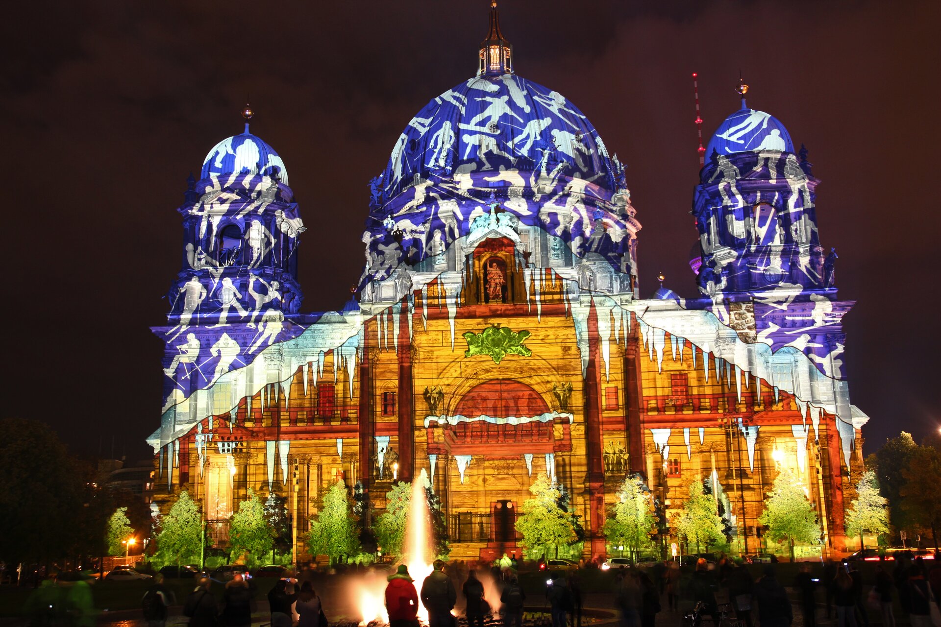 Ilustracja przedstawia budynek w Berlinie, na którym wyświetlany jest 3D mapping. W górnej części, na niebieskim tle ukazane są białe postacie podczas jazdy na nartach oraz snowboardzie [czyt. snołbordzie]. Dolna część przedstawia wyświetloną fasadę typu bazylikowego z dekoracyjnym, półokrągłym wejściem. Przed budynkiem rosną choinki.