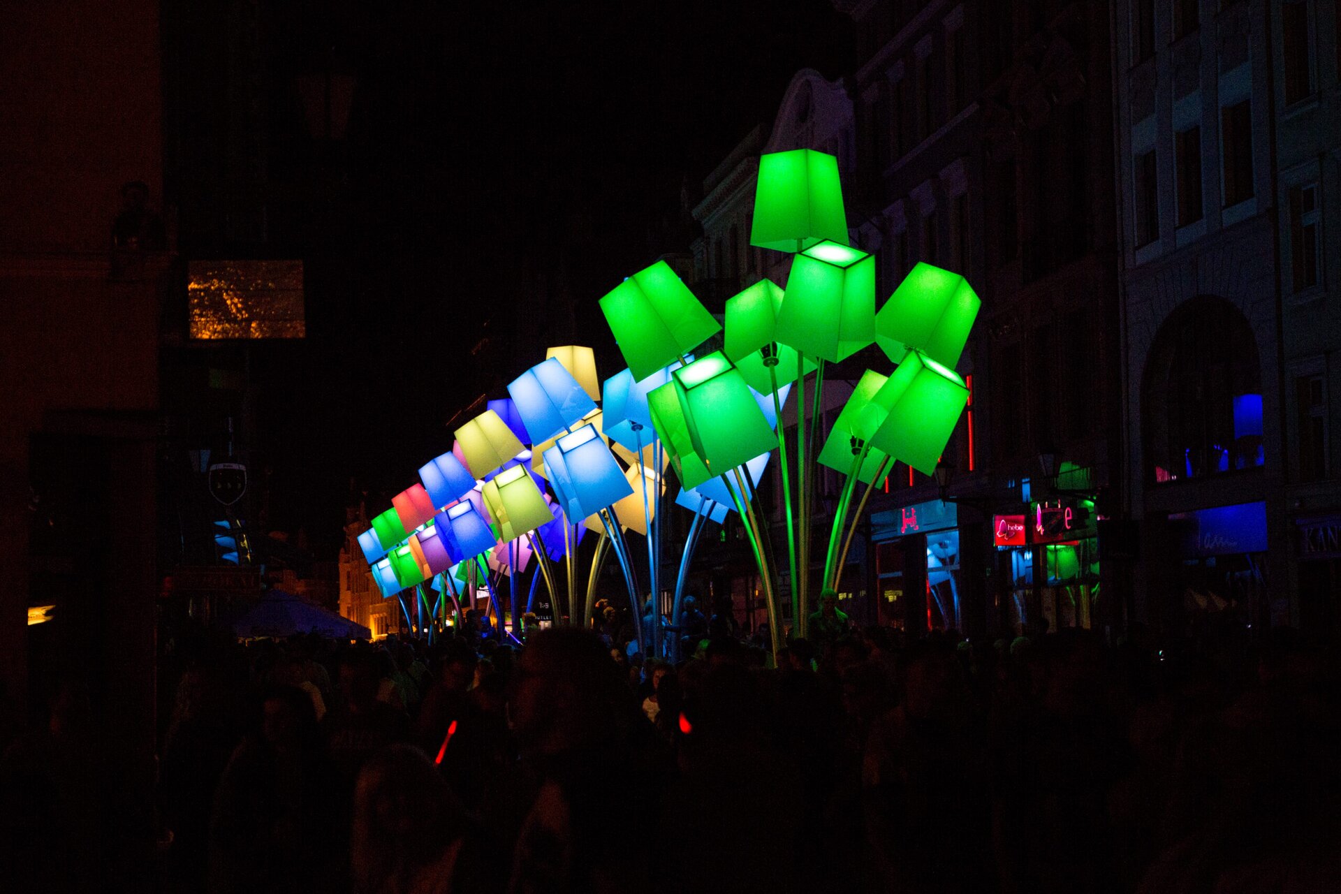 Na zdjęciu znajduje się duża instalacja w następujących kolorach: żółtym, zielonym, niebieskim, granatowym, fioletowym, różowym.  Przedstawia stojące lampy. Dokoła niej znajduje się tłum ludzi, a w tle są budynki. Fotografia jest ciemna, jedynie instalacja jest rozświetlona.