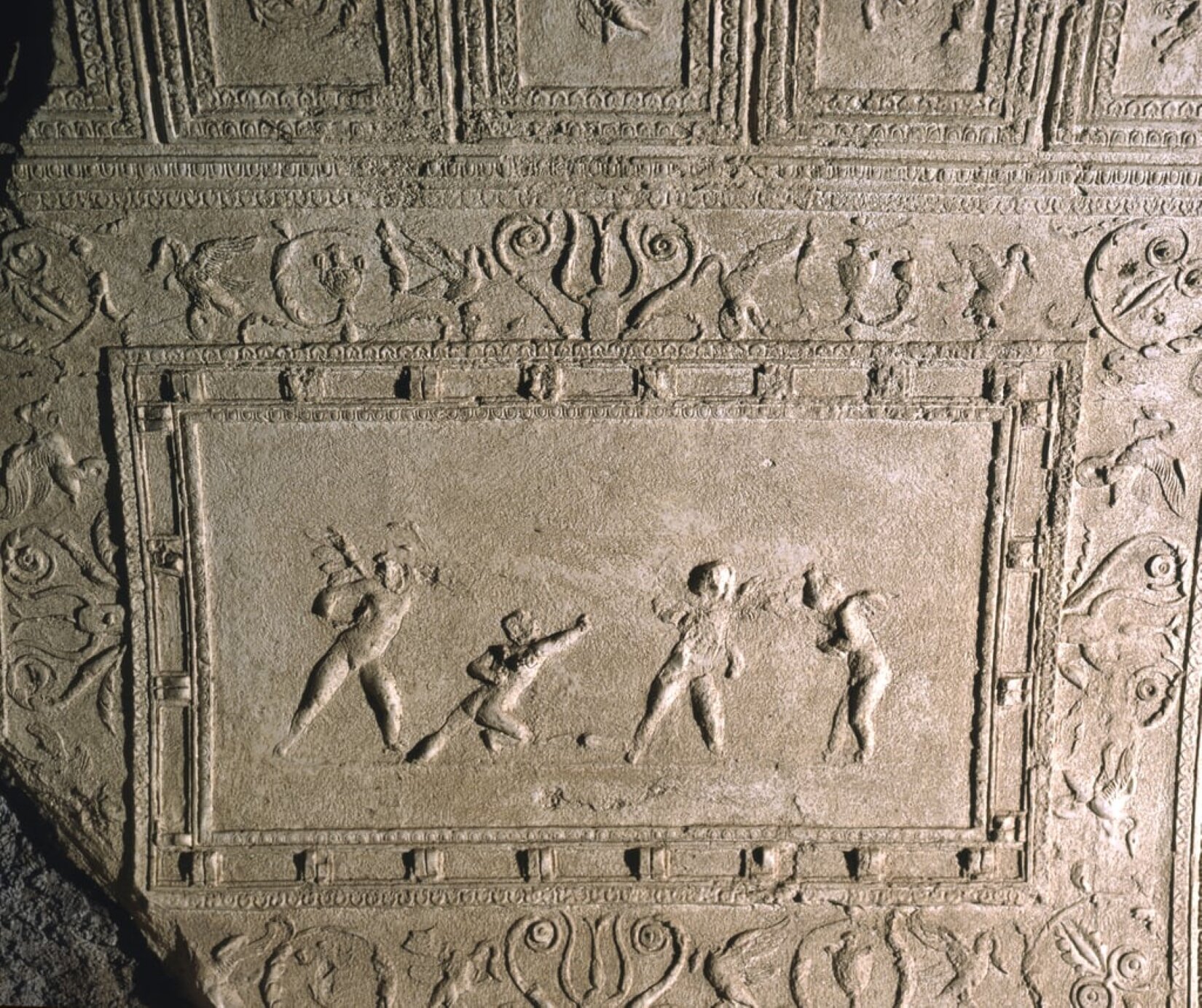 Fotografia przedstawia płaskorzeźbę na prostokątnym fragmencie muru. Ukazane są cztery postaci Amorów w różnych pozach. Całość otoczona jest obramowaniem; nad nim znajdują się liściaste ornamenty. Praca utrzymana jest w kolorach szarości.