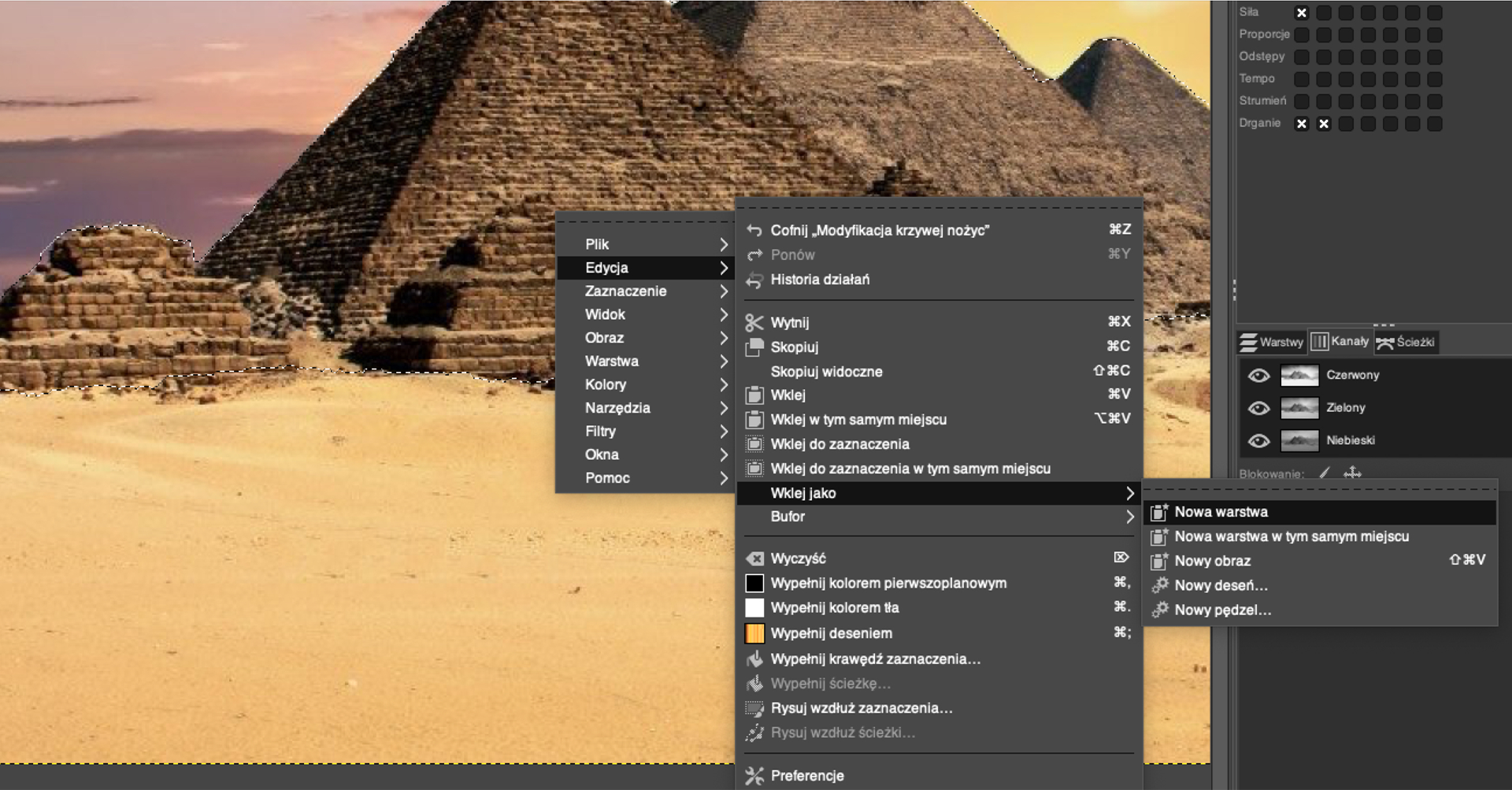 Ilustracja przedstawia okno w programie GIMP. Wewnątrz okna znajduje się fragment zdjęcia z piramidami – podstawy piramid i piaszczysta pustynia. Na tle obrazka znajduje się pionowa lista z pozycjami: Plik, Edycja, Zaznaczenie, Widok, Obraz, Warstwa, Kolory, Narzędzia, Filtry, Okna, Pomoc. Zaznaczona jest opcja Edycja, a obok niej rozwinięta kolejna pionowa lista z pozycjami: Cofnij „Modyfikacja krzywej nożyc”, Ponów, Historia działań, Wytnij, Skopiuj, Skopiuj widoczne, Wklej, Wklej w tym samym miejscu, Wklej do zaznaczenia, Wklej do zaznaczenia w tym samym miejscu, Wklej jako, Bufor, Wyczysć, Wypełnij kolorem pierwszoplanowym, Wypełnij kolorem tła, Wypełnij deseniem, Wypełnij krawędź zaznaczenia…, Wypełnij ścieżkę, Rysuj wzdłuż zaznaczenia…, Rysuj wzdłuż ścieżki…, Preferencje. Z listy wybrana jest pozycja Wklej jako, obok rozwinięta kolejna lista z pozycjami: Nowa warstwa, Nowa warstwa w tym samym miejscu, Nowy obraz, Nowy deseń…, Nowy pędzel. Z listy wybrano pozycję: Nowa warstwa. Z prawej strony znajduje się pasek menu. 