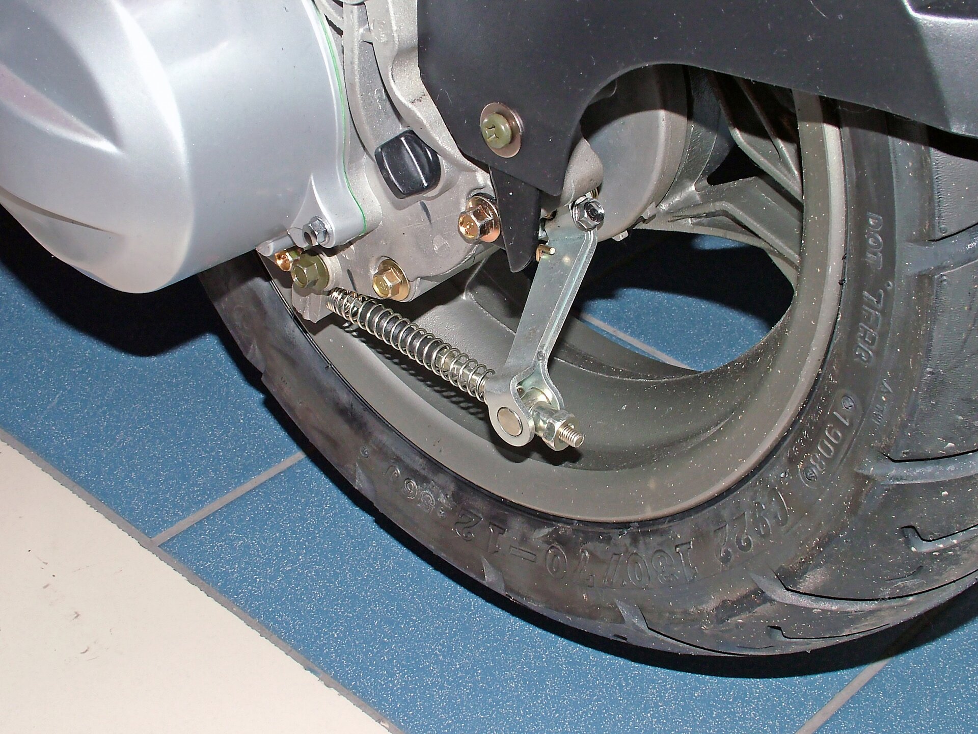 Na ekranie opona pojazdu ze zbliżeniem na korek wlewowy i śruba spustowa oleju smarującego kaskadę kół zębatych stanowiących końcową przekładnię skutera.