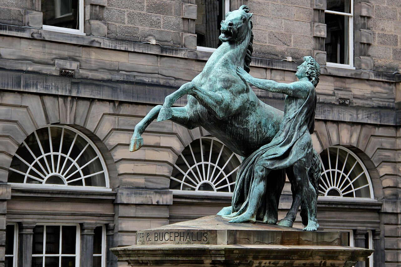 Zdjęcie przedstawia pomnik. Ukazany jest koń stojący na tylnych kopytach i próbujący go uspokoić młody mężczyzna w powłóczystej szacie sięgającej ziemi.