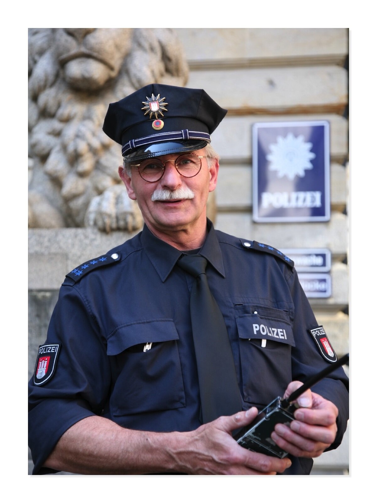 Fotografia przedstawia mężczyznę, który wykonuje zawód policjanta. Ubrany jest w mundur, ma na głowie policyjną czapkę, w dłoni trzyma krótkofalówkę. Za nim widoczna jest komenda policji.