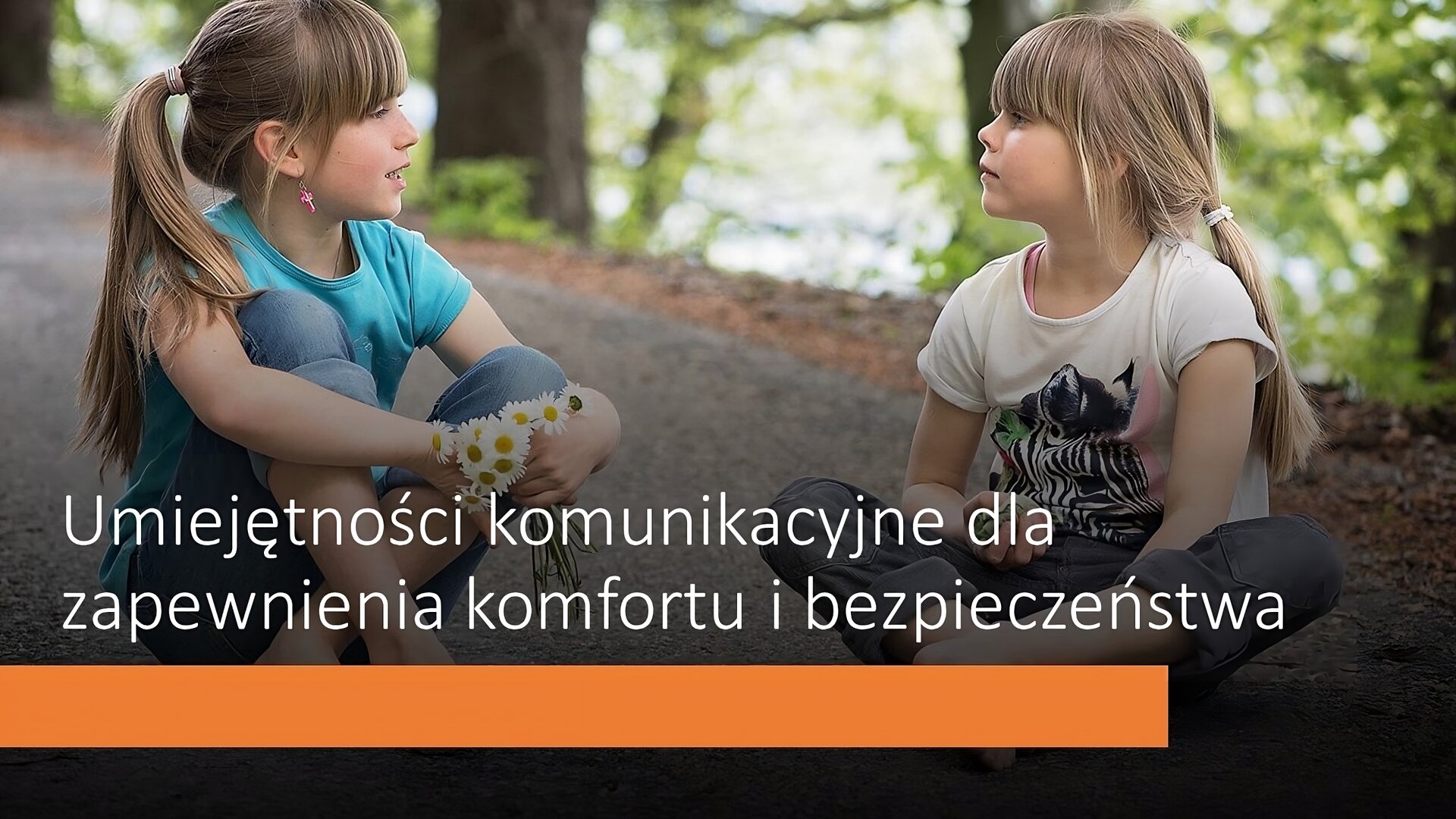 Slajd, na którym znajduje się zdjęcie dwóch dziewczynek. Siedzą po turecku i wpatrują się w siebie. Obie mają długie, blond włosy upięte w kitkę oraz grzywkę. Mają na sobie koszulki z krótkim rękawkiem: niebieską oraz białą. Dziewczynka siedząca po lewej stronie trzyma w dłoni bukiet stokrotek. W tle piaskowa droga i zielone drzewa. U dołu slajdu tekst: „Umiejętności komunikacyjne dla zapewnienia komfortu i bezpieczeństwa”, a pod nim pomarańczowy pasek.