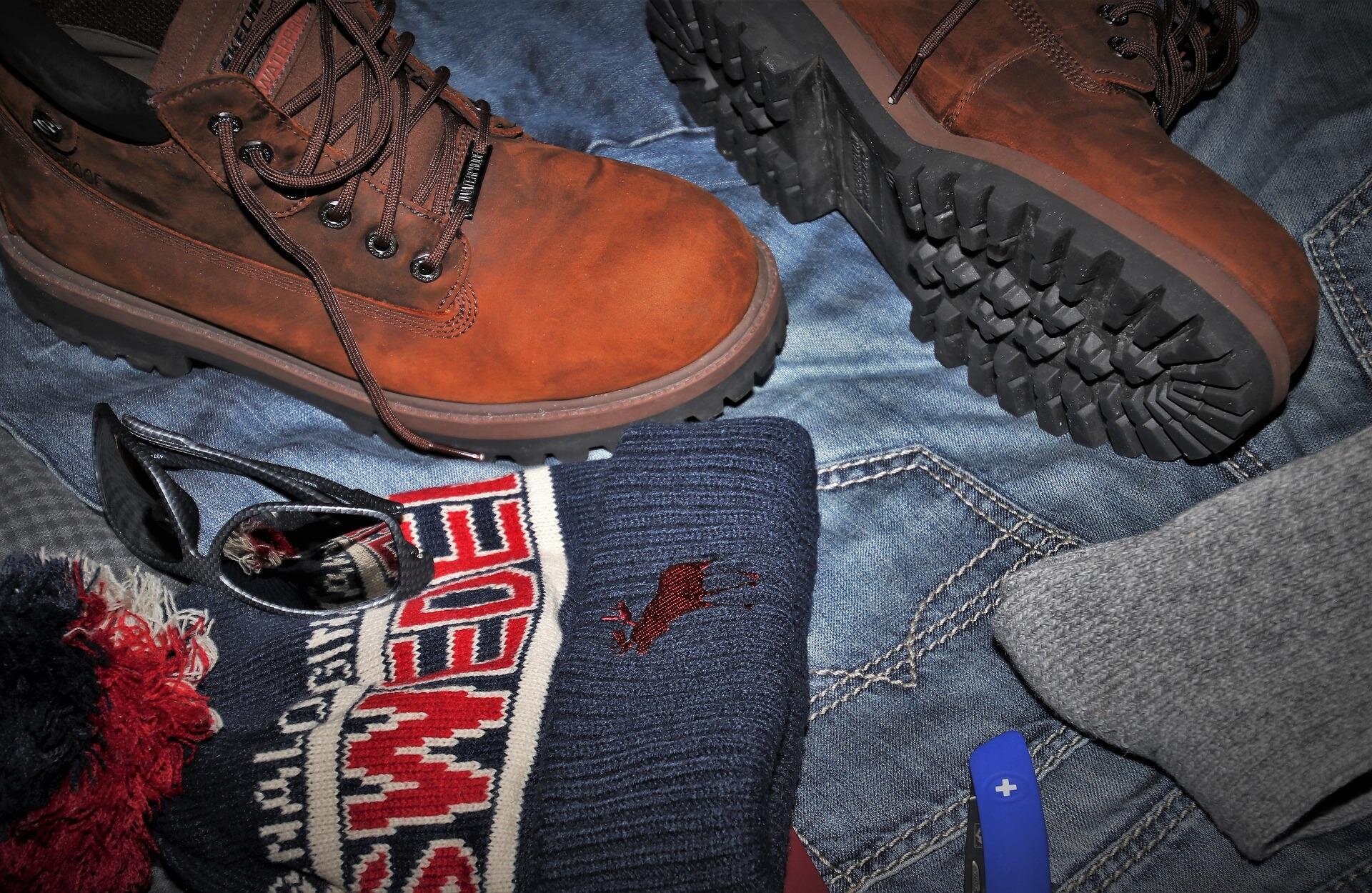 Zdjęcie przedstawia górskie buty, okulary przeciwsłoneczne, dżinsy i ciepłką czapkę