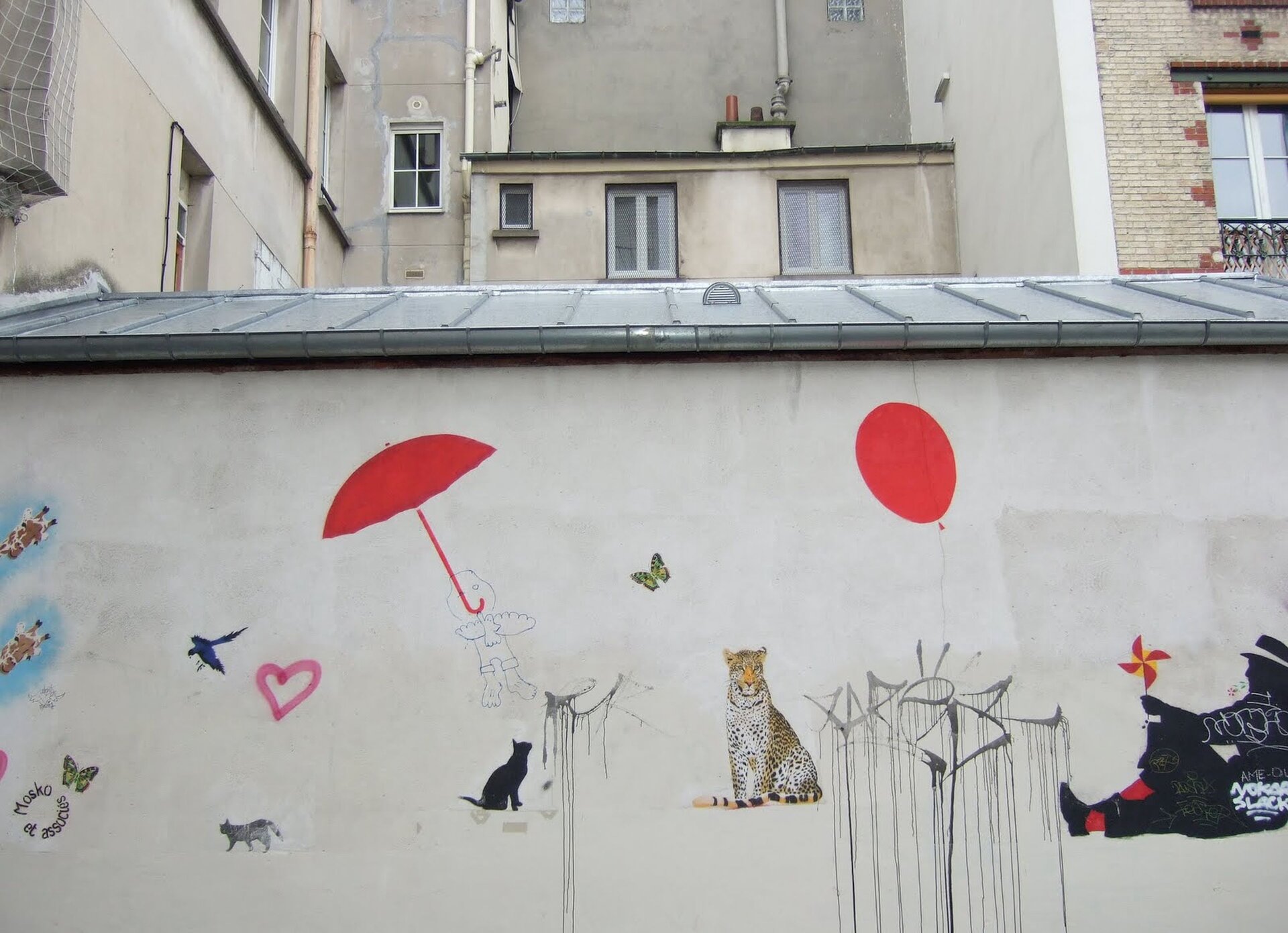 Ilustracja przestawia mural Mosko et Associes. Ukazuje namalowane na ścianie, swobodnie rozmieszczone: czerwone parasol i balon, czarnego kota, tygrysa, ptaki, motyla i siedzącego mężczyznę. Za murem znajdują sie budynki.
