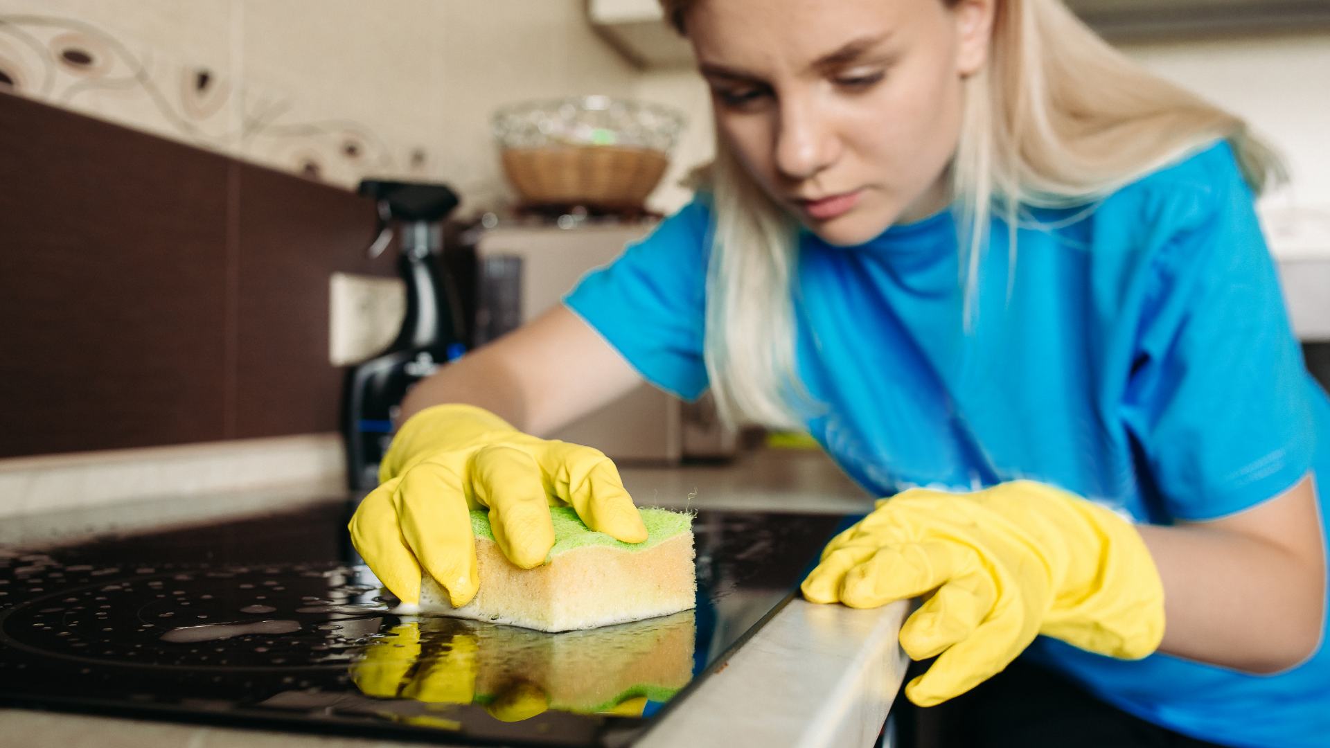 Zdjęcie przedstawia młodą kobietę, która czyści kuchnię. Na rękach ma gumowe rękawice oraz gąbkę w dłoni.