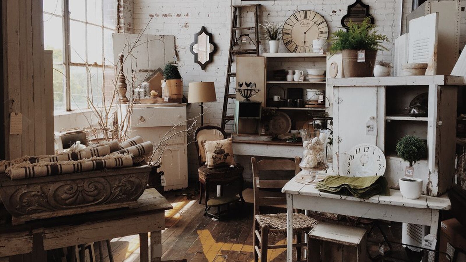 Zdjęcie przedstawia pomieszczenie wypełnione różnymi antycznymi meblami i przedmiotami. Widoczne są białe kredensy, szafki z szufladami, krzesło z podnóżkiem, krzesło z wyściełanym siedziskiem, stojące na półkach i wiszące na ścianach zegary, lampy, tkaniny, naczynia, poduszki.