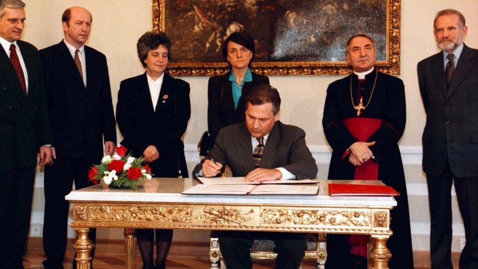 Zdjęcie przedstawia Prezydenta Aleksandra Kwaśniewskiego, podpisującego dokument konkordatu. W jego otoczeniu stoi trzech mężczyzn w garniturach, biskup i dwie kobiety.