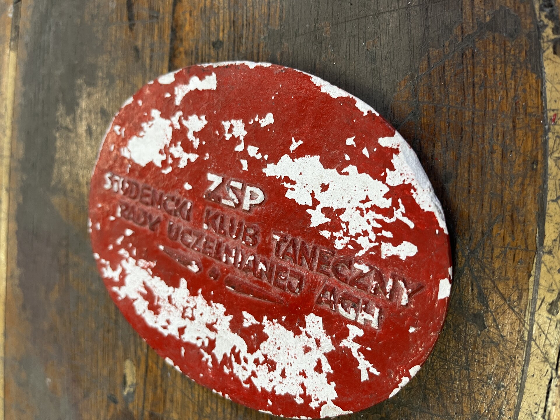 Na zdjęciu przedstawiono drewniany model odlewniczy z widocznymi brakami. Przedstawiony model jest w kształcie okręgu. W centralnej części modelu znajduje się napis: ZSP Studencki klub taneczny Rady Uczelnianej AGH. Na całej powierzchni modelu widoczne są ubytki farby. Ubytki są w kolorze białym