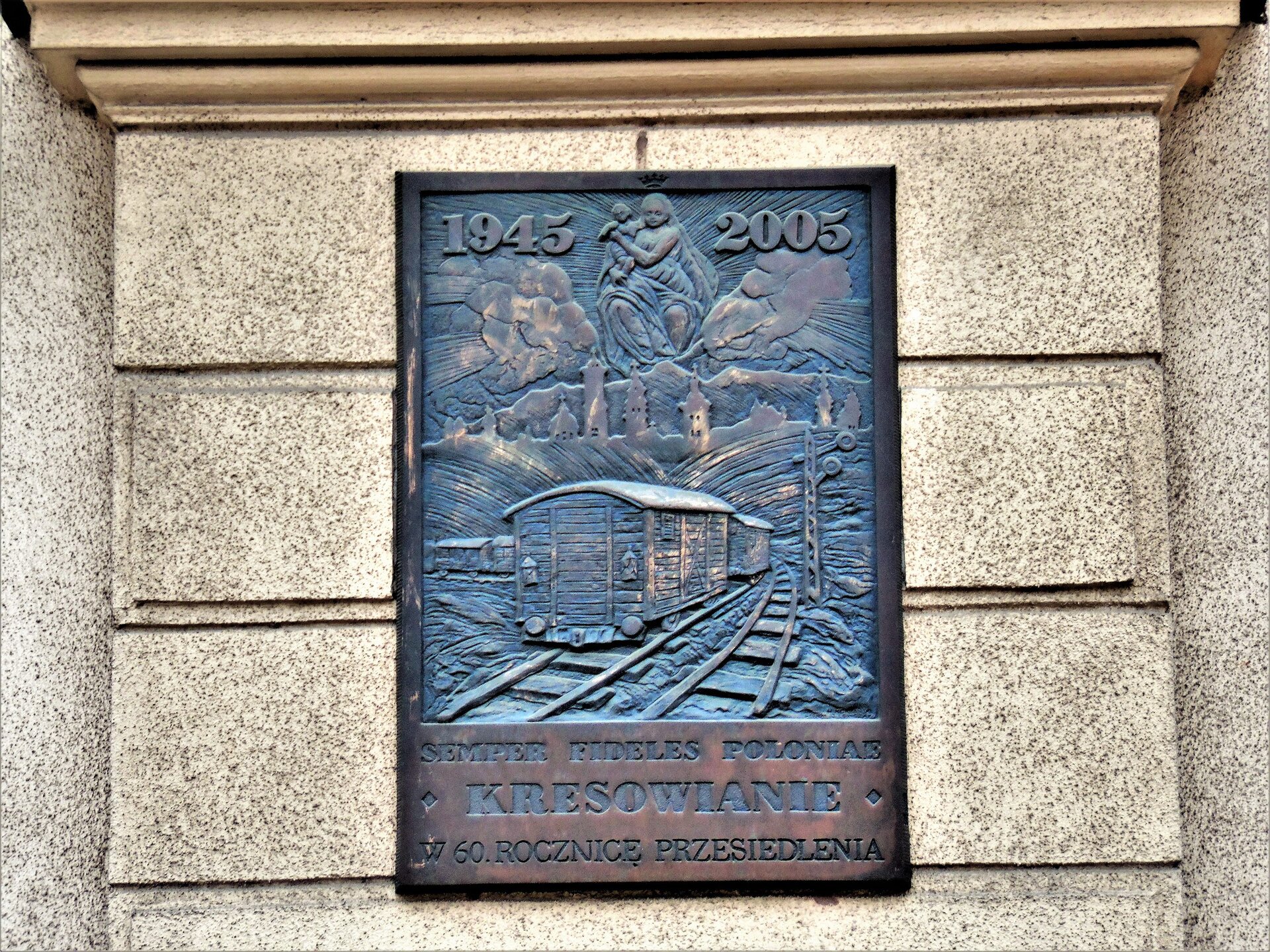 Zdjęcie przedstawia pamiątkową tablicę, umieszczoną na elewacji budynku. Na tablicy widać tył wagonu odjeżdżającego pociągu towarowego. W tle pagórki, nad nimi chmury i wyłaniająca się zza nich Matka Boska z dzieciątkiem. Na górze data 1945 oraz 2005. Na dole napis po łacinie: "Semper fidelis Poloniae". Pod nim: "Kresowianie w 60 rocznicę przesiedlenia".