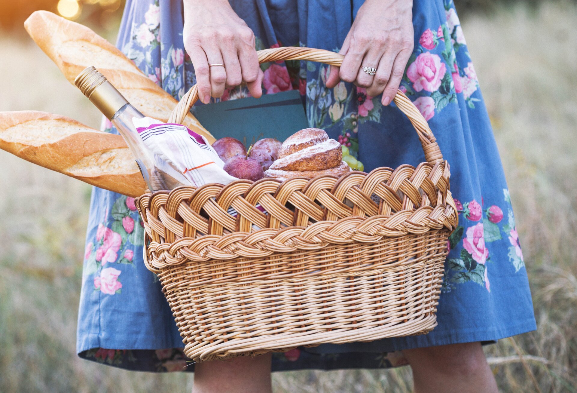 Zdjęcie kosza o owalnym dnie z ozdobnym wykończeniem na górze i uchwytem. W koszu znajdują się bagietki, pieczywo, butelka wina i owoce. Trzymany jest przez kobietę, której postać widoczna jest tylko w połowie.