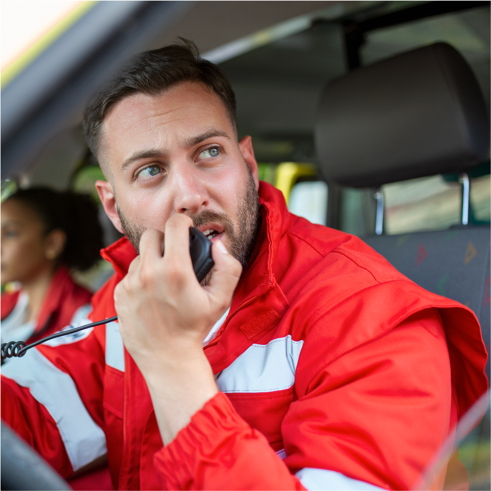 Zdjęcie przedstawia ratownika medycznego w czerwonej kurtce, który siedzi za kierownicą karetki i rozmawia przez radiostację.